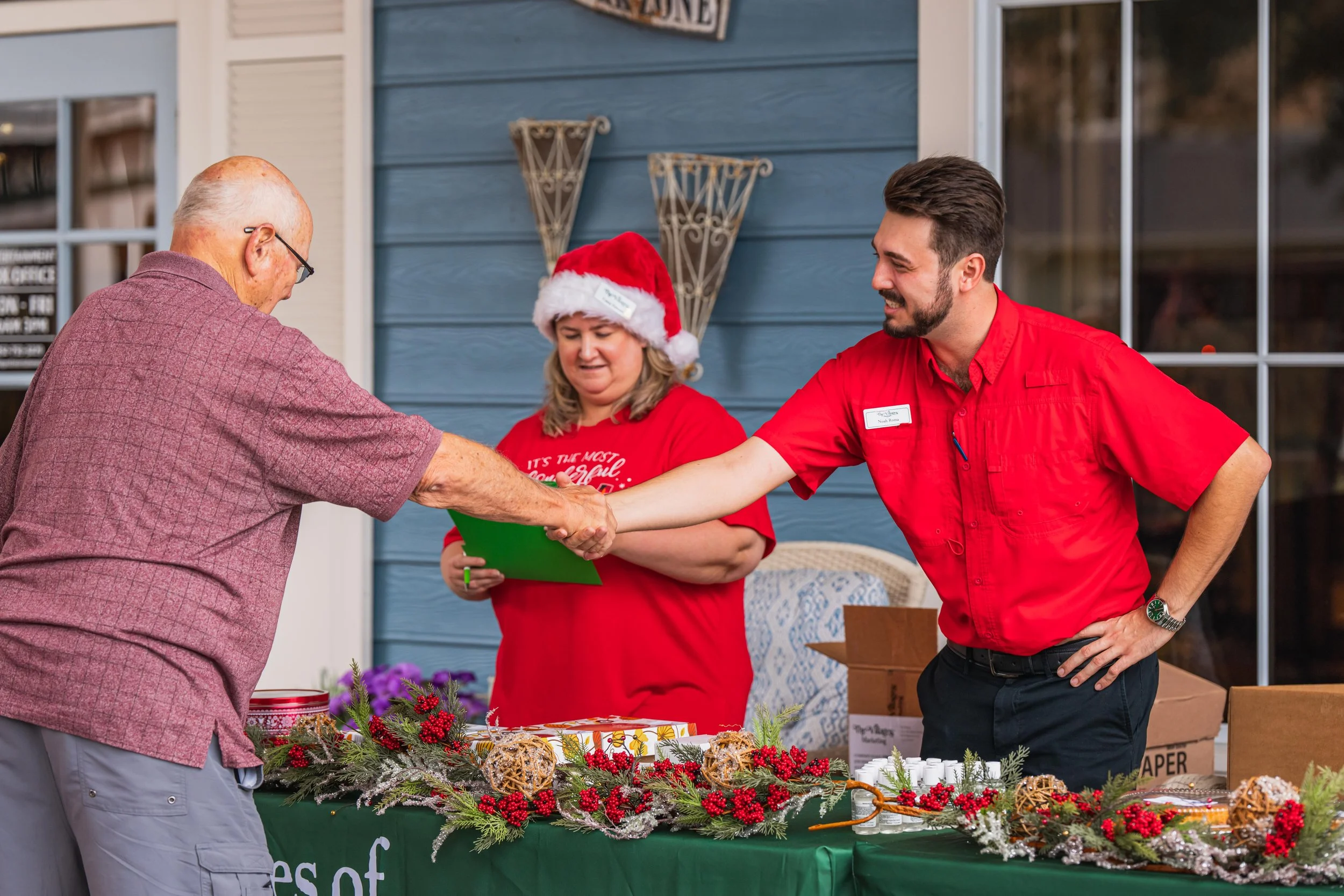 An elderly man shaking hands with a young man in front of a decorated table at a holiday event, with a woman in a Santa hat standing behind them.
