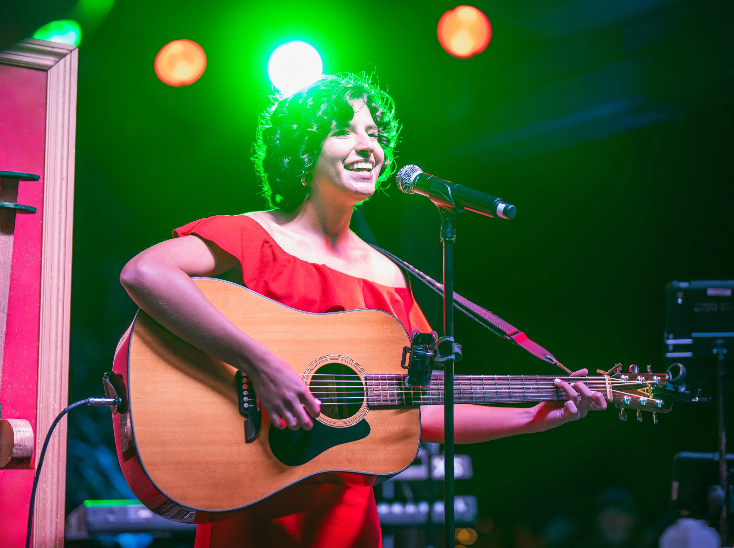 Christina Vukel playing an acoustic guitar and singing into a microphone on stage, illuminated by colorful stage lights.