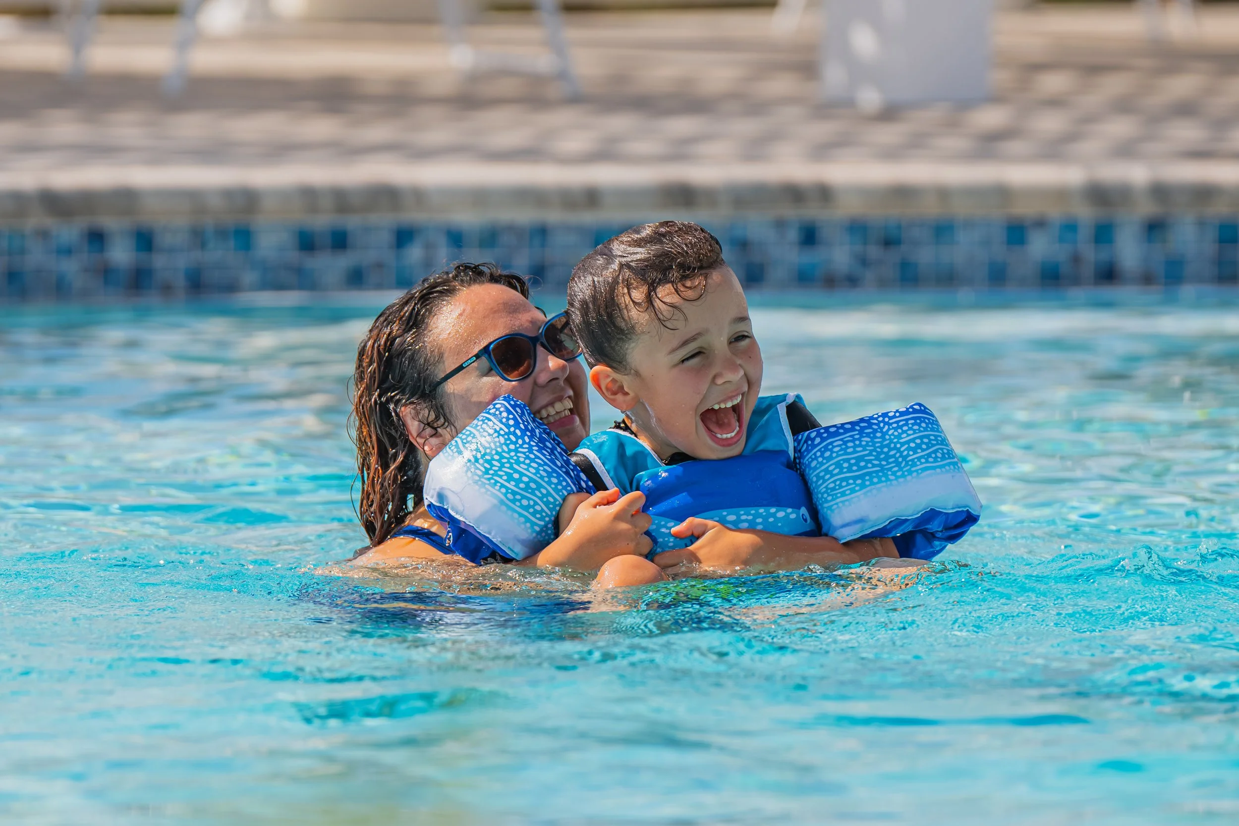 A woman and a young boy smiling and enjoying themselves while swimming in a pool, wearing blue floaties.