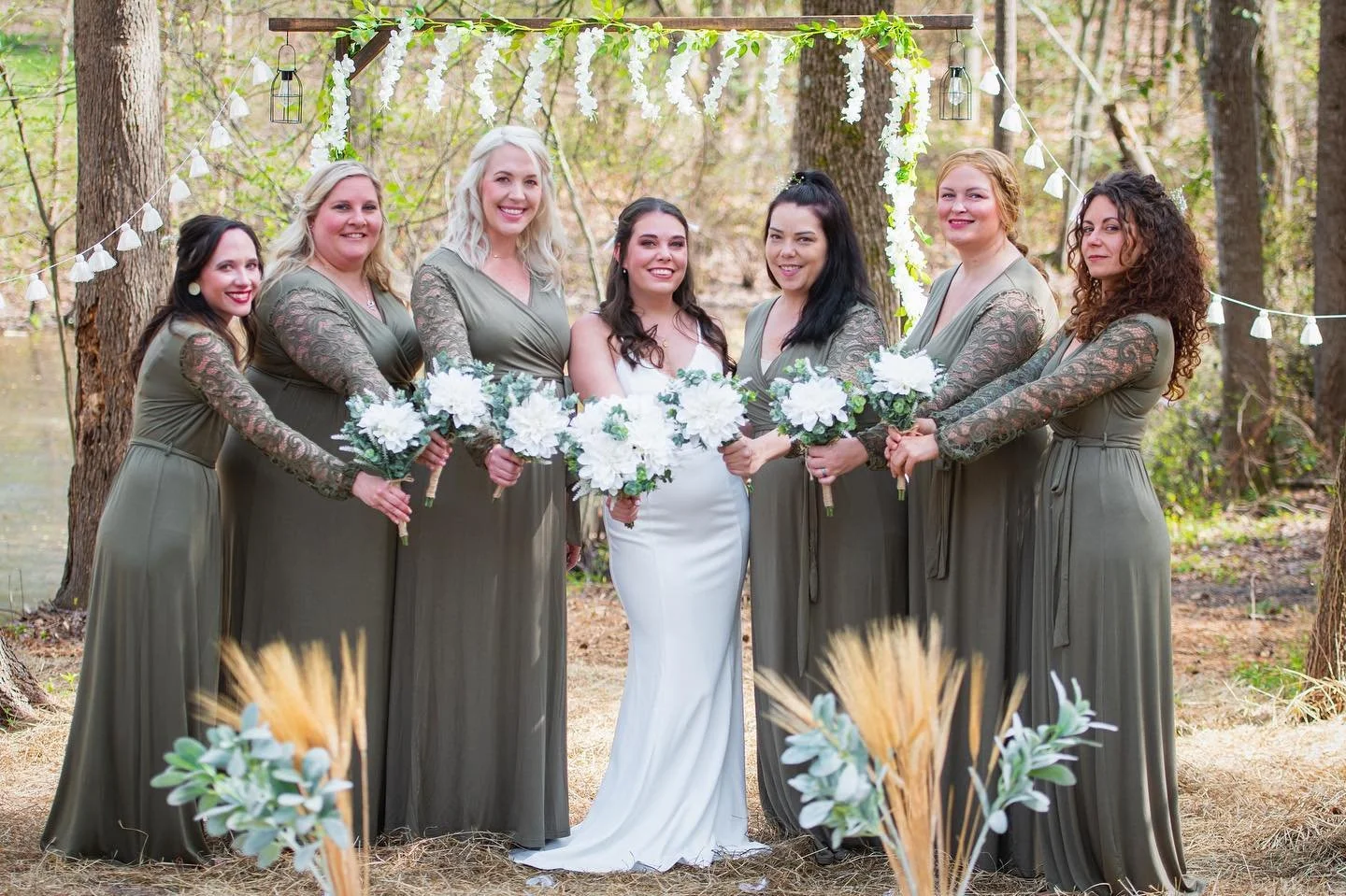 Bridal party at a wedding in a wooded outdoor setting, with seven women in coordinated olive green dresses, holding white floral bouquets, under a decorative arch with hanging lights and floral accents.