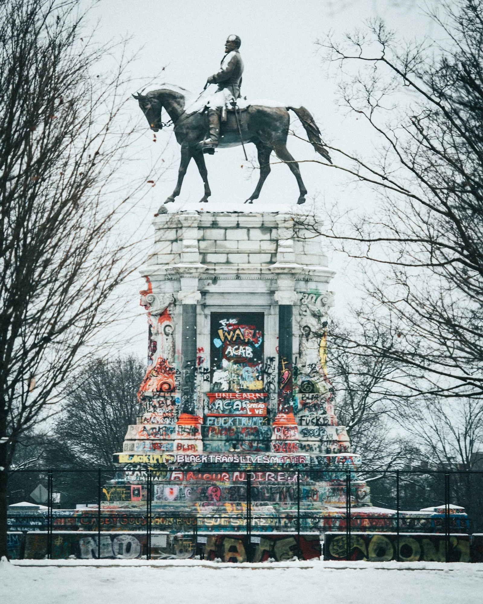 An equestrian statue on a large, graffiti-covered pedestal with a snow-covered ground and leafless trees in the background.
