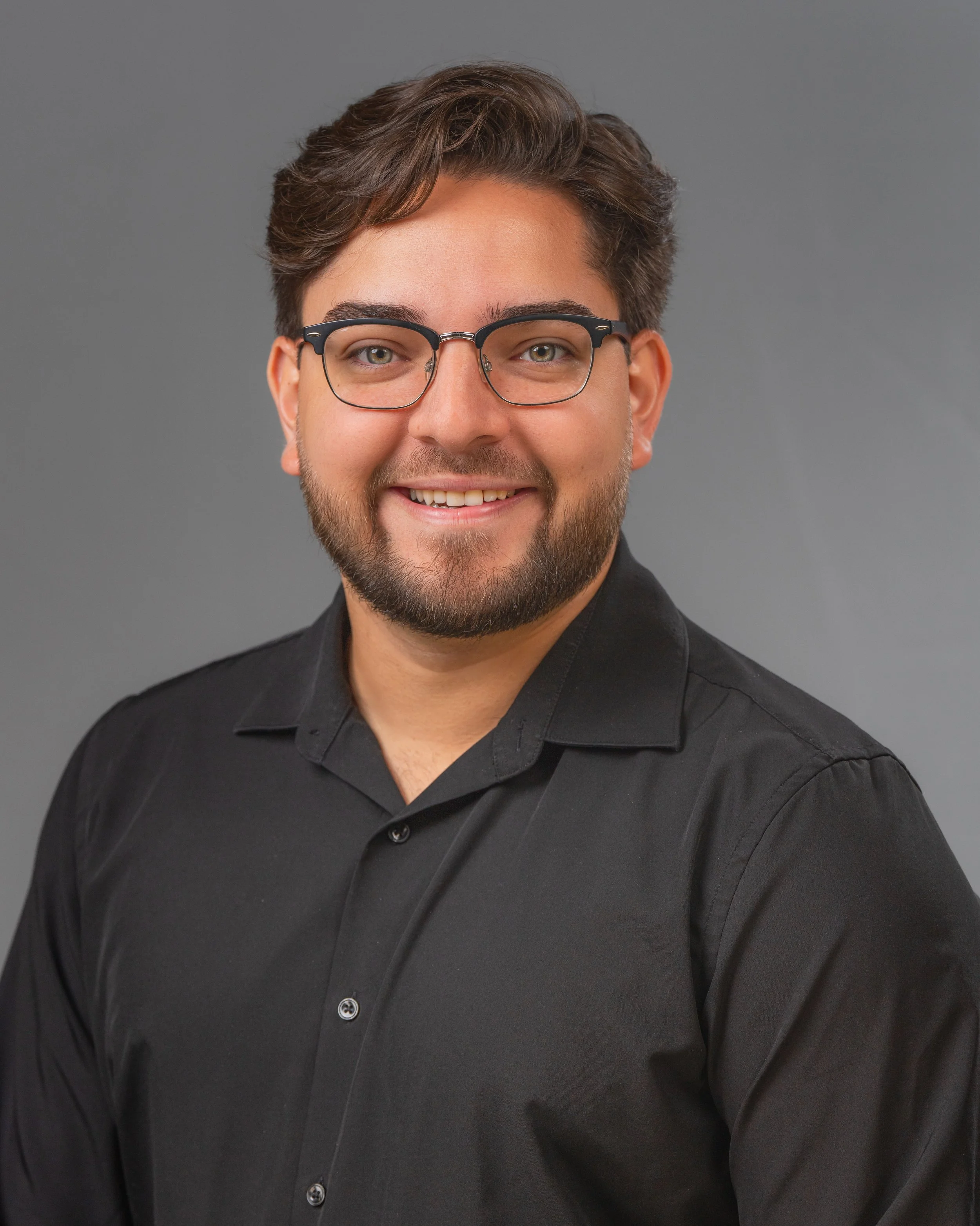 Portrait photography, Central Florida. Corporate headshots. A young man with glasses and a beard smiling, wearing a black shirt against a gray background.