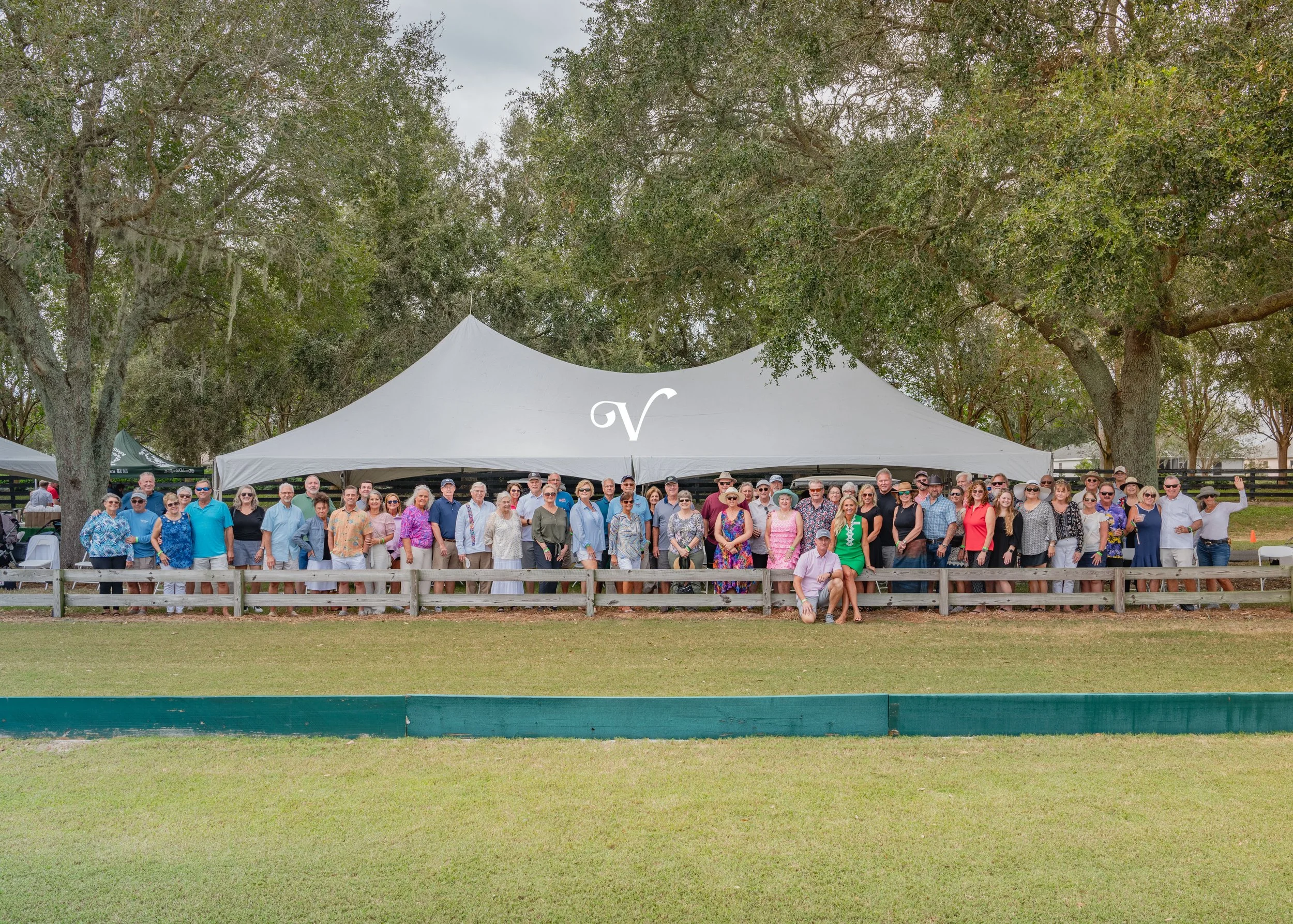 Group of people standing under a large white tent outdoors, posing for a photo with trees in the background.