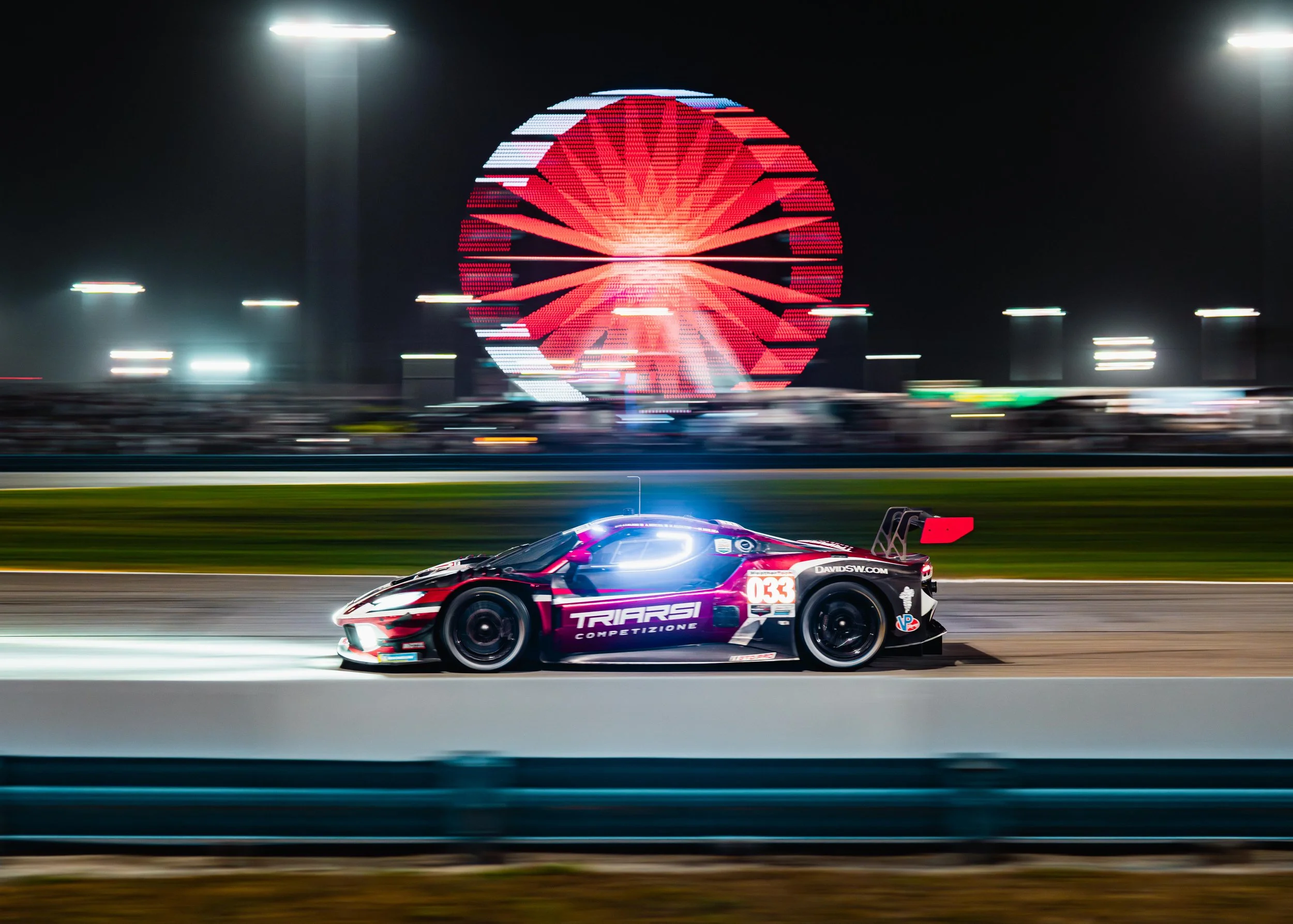 Nighttime race car on track with a bright red Ferris wheel in the background, motion blur effect.