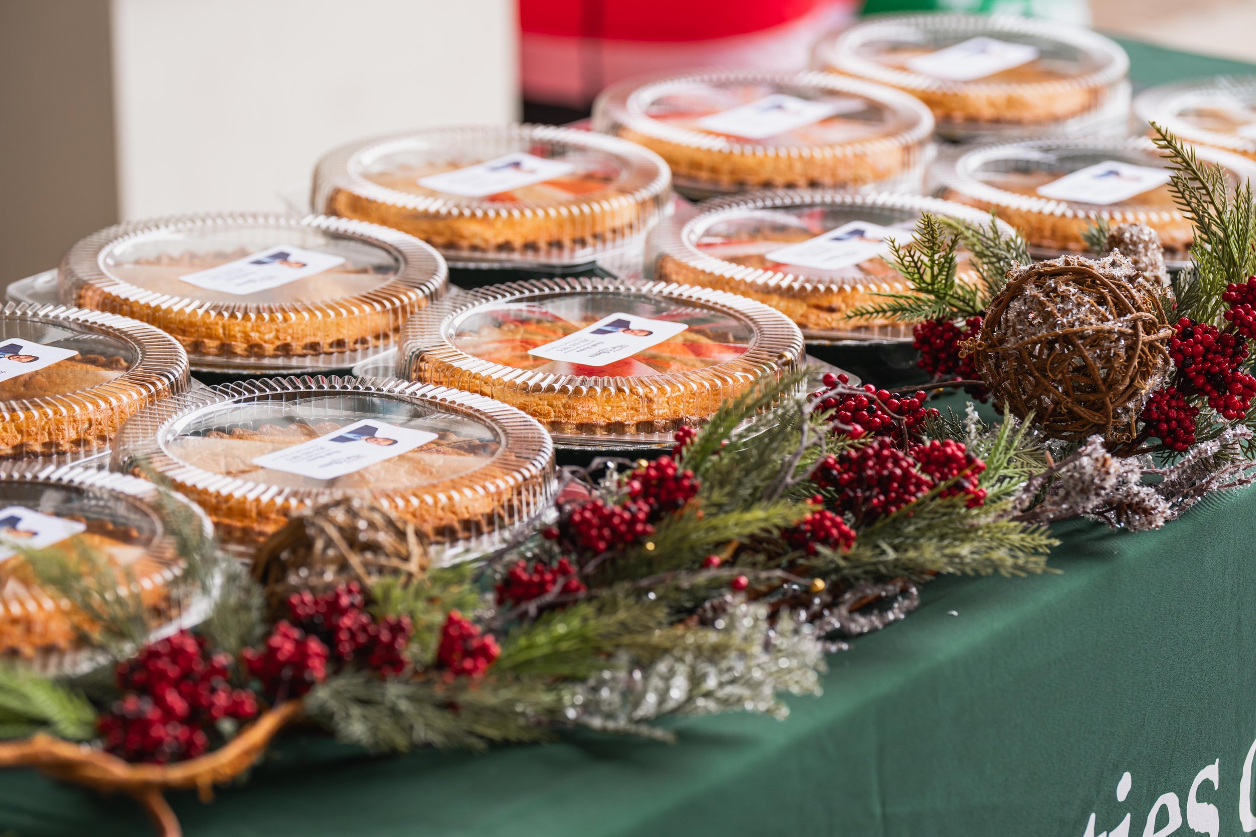Several clear plastic containers of pie on a decorated table with holiday greenery and red berries.