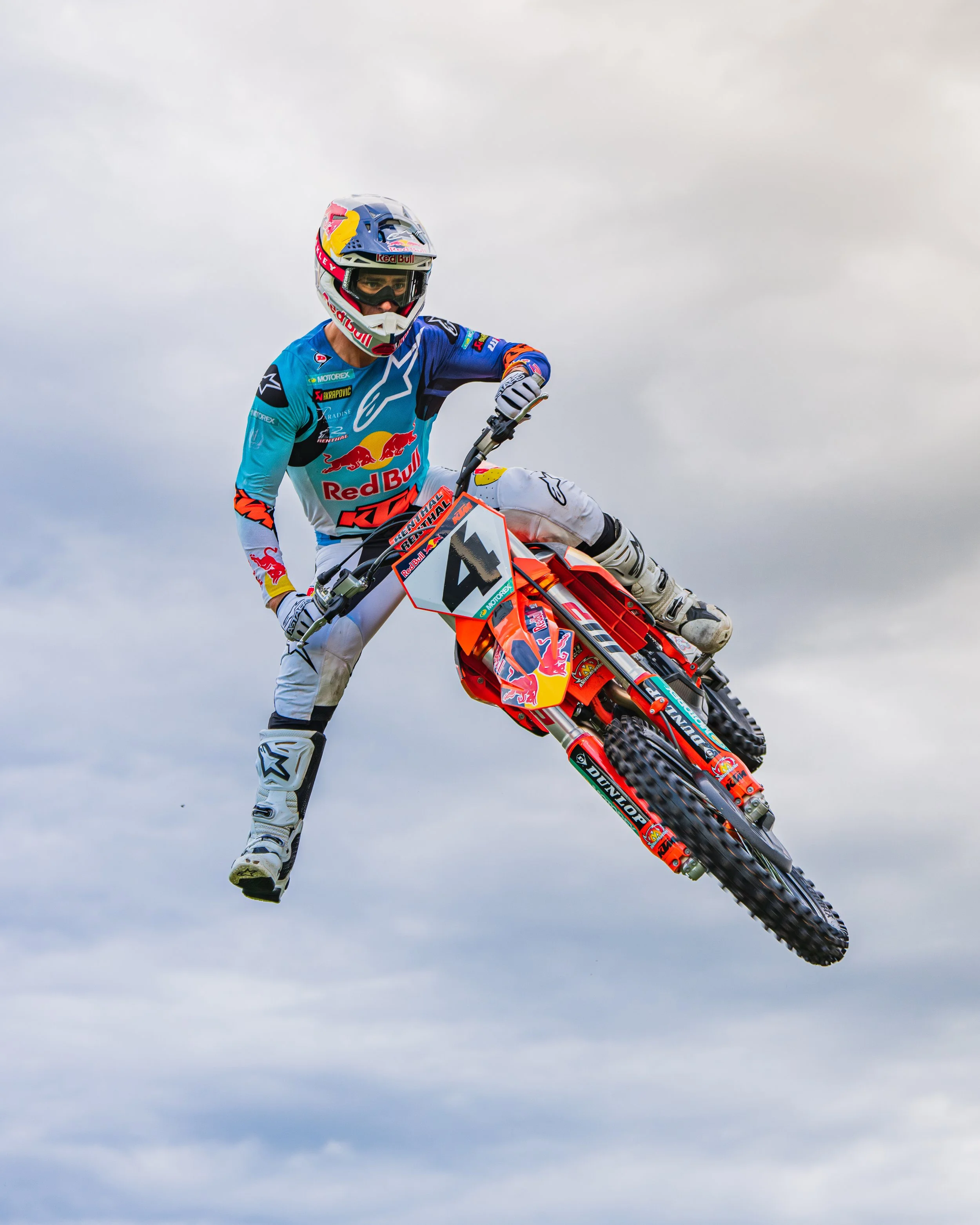Sports photography. Action Sports Motocross rider wearing a white helmet, blue and white racing gear, riding a red dirt bike in mid-air against cloudy sky background. Chase Sexton Red Bull