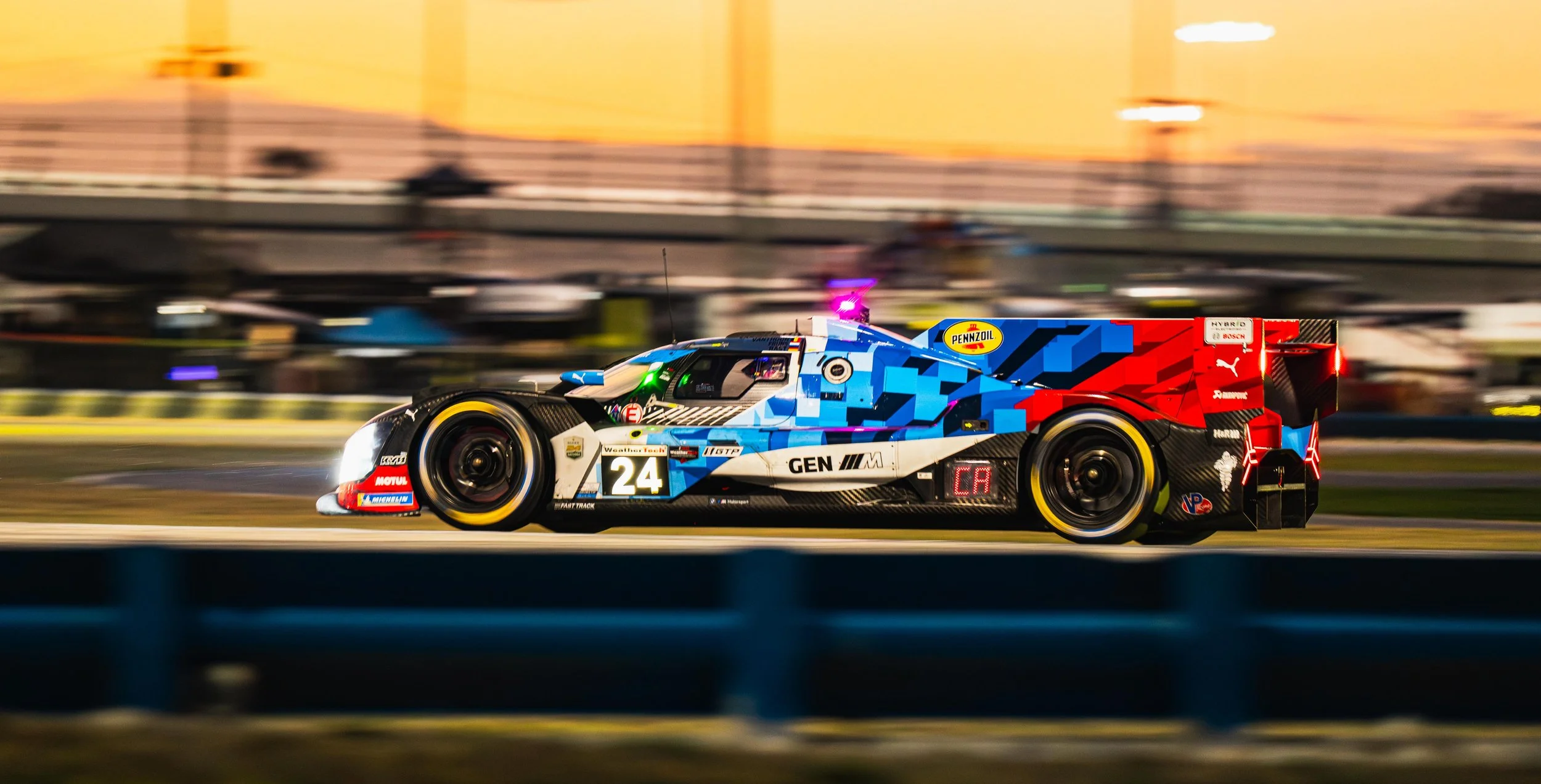 A race car speeding on a track at sunset, painted in blue, black, white, red, and yellow, with sponsor logos including Pennzoil, Motul, Michelin, and Puma.