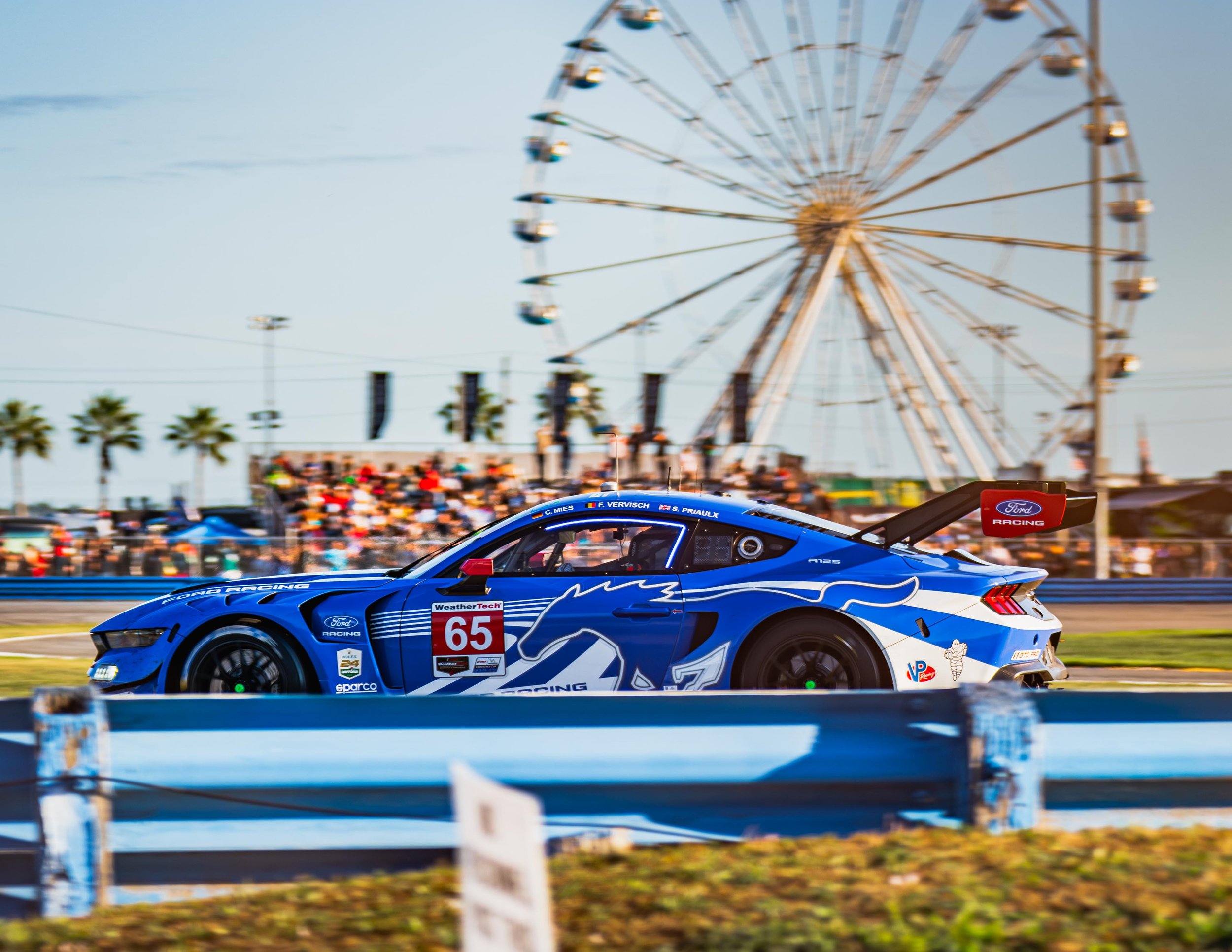 A blue race car with Sponsor logos, including a unicorn graphic, racing on a track with a Ferris wheel and spectators in the background.