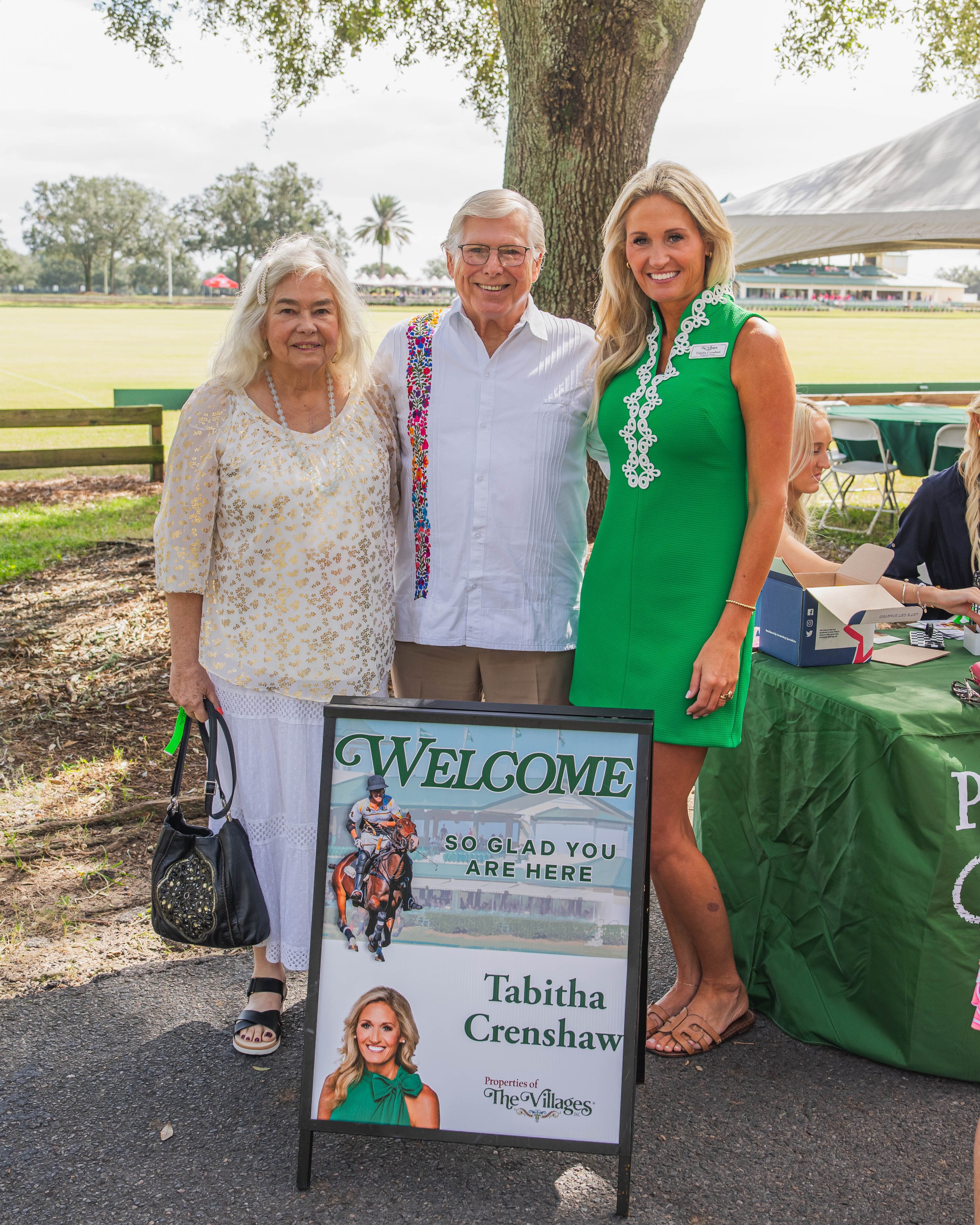 Three people standing outdoors next to a welcome sign for Tabitha Crenshaw at an event, with a grassy field and trees in the background.