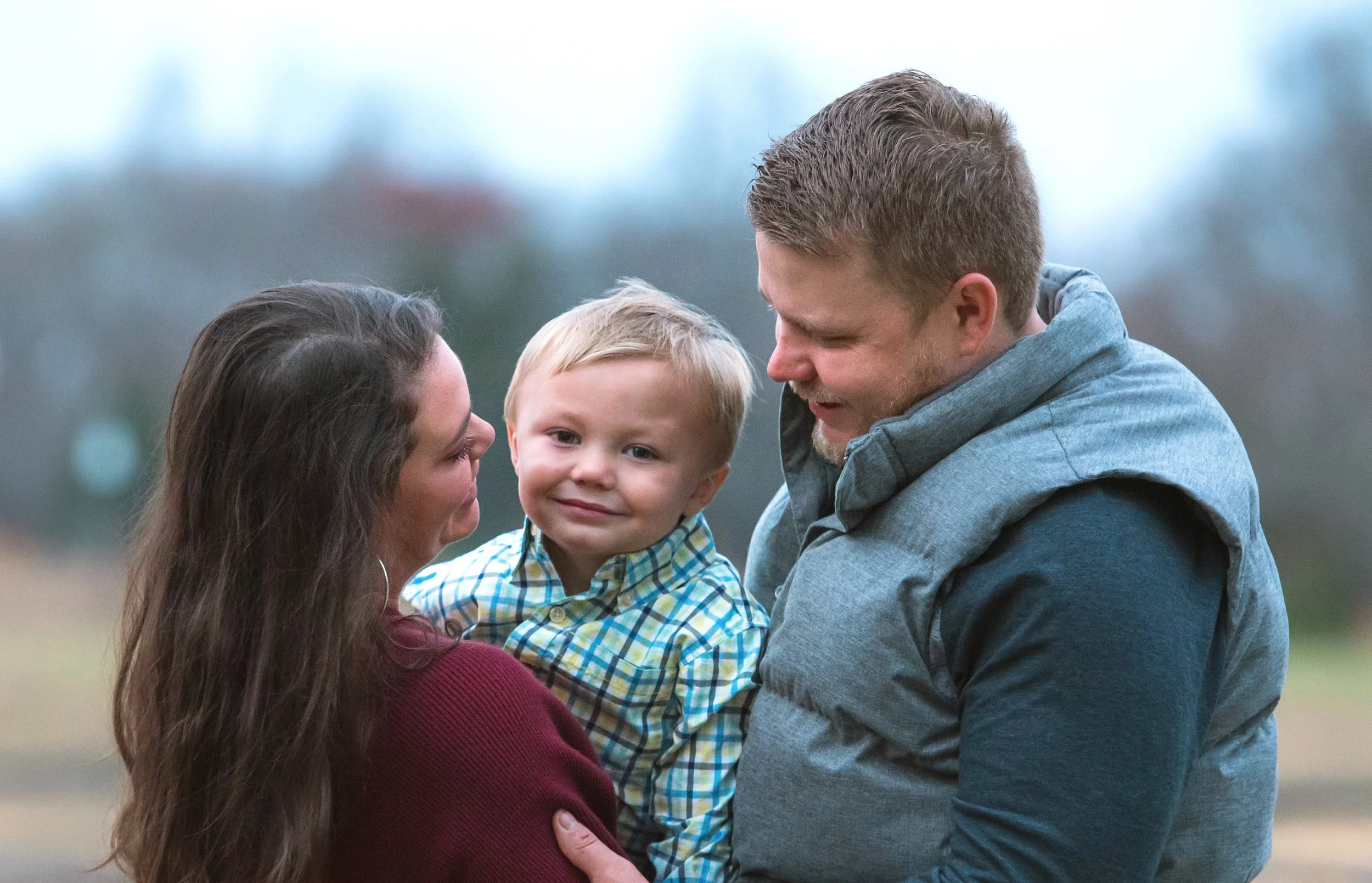 A family of three outdoors, a woman holding a young boy and a man looking at the boy, with a blurred natural background.