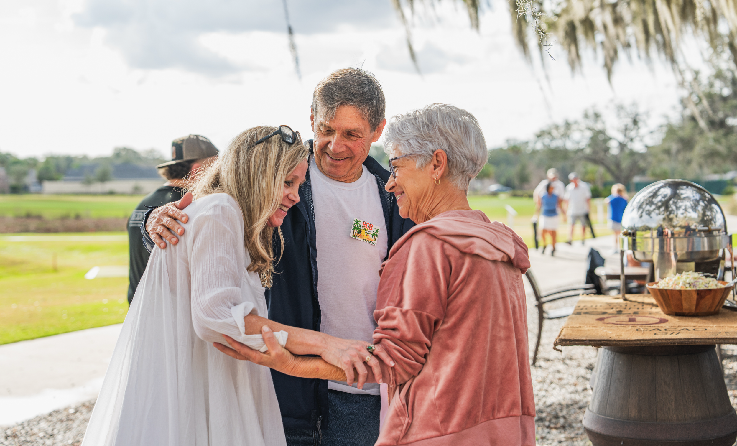 Three people smiling and hugging each other outdoors at a gathering, with a buffet table on the right and a park in the background