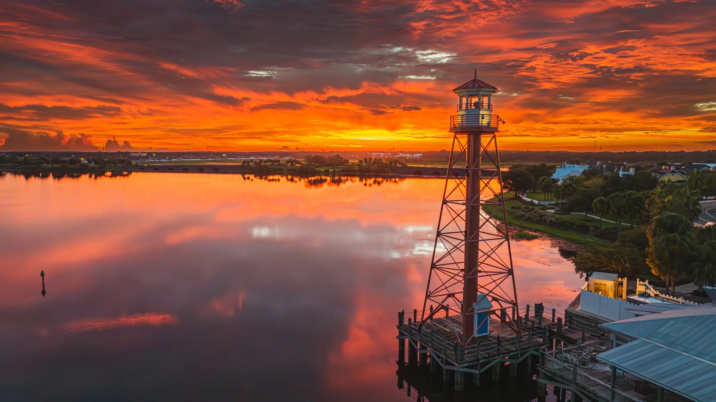 A scenic sunrise over Lake Sumter Landing with a tall lighthouse on a pier in the foreground. The sky is filled with vibrant orange and pink clouds, reflecting beautifully on the water.