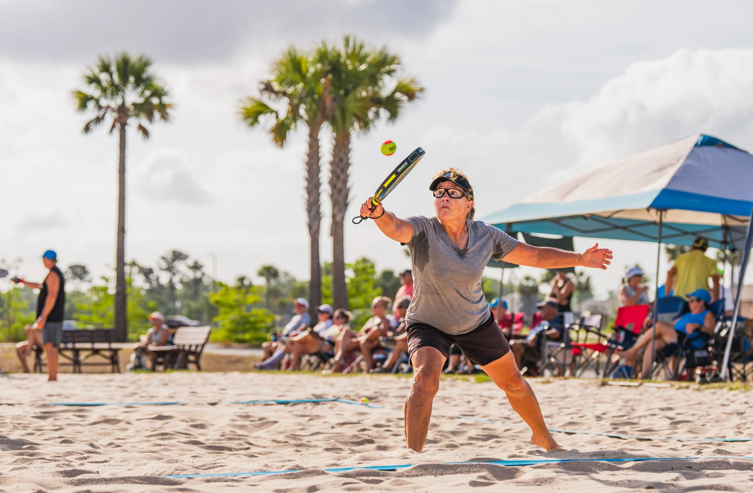 Older woman playing pickleball on a sandy court with palm trees and spectators in the background.