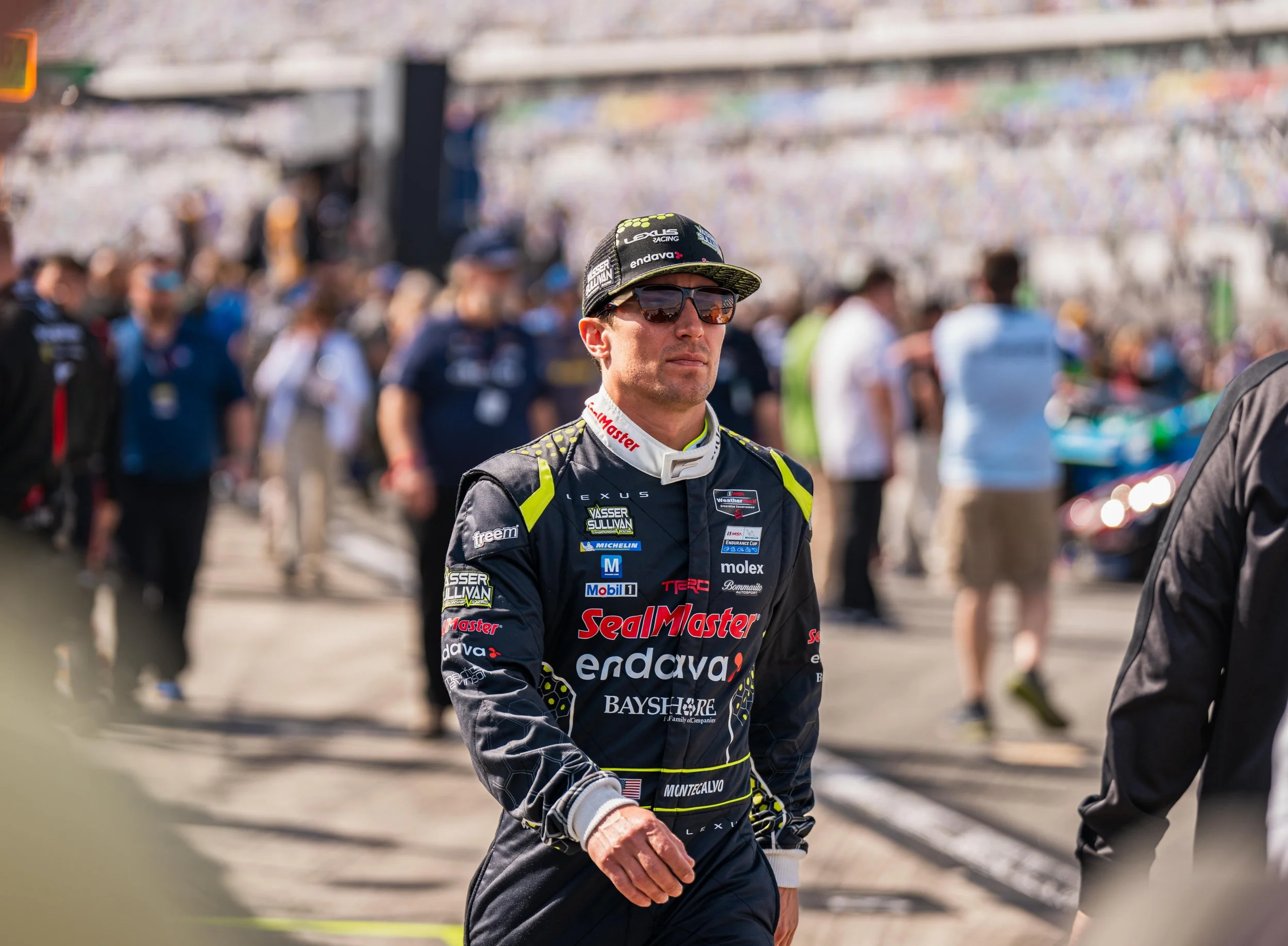 A race car driver walking in the pit lane during a racing event, wearing a black racing suit with various sponsor logos and a black cap with logos, sunglasses, and a focused expression.