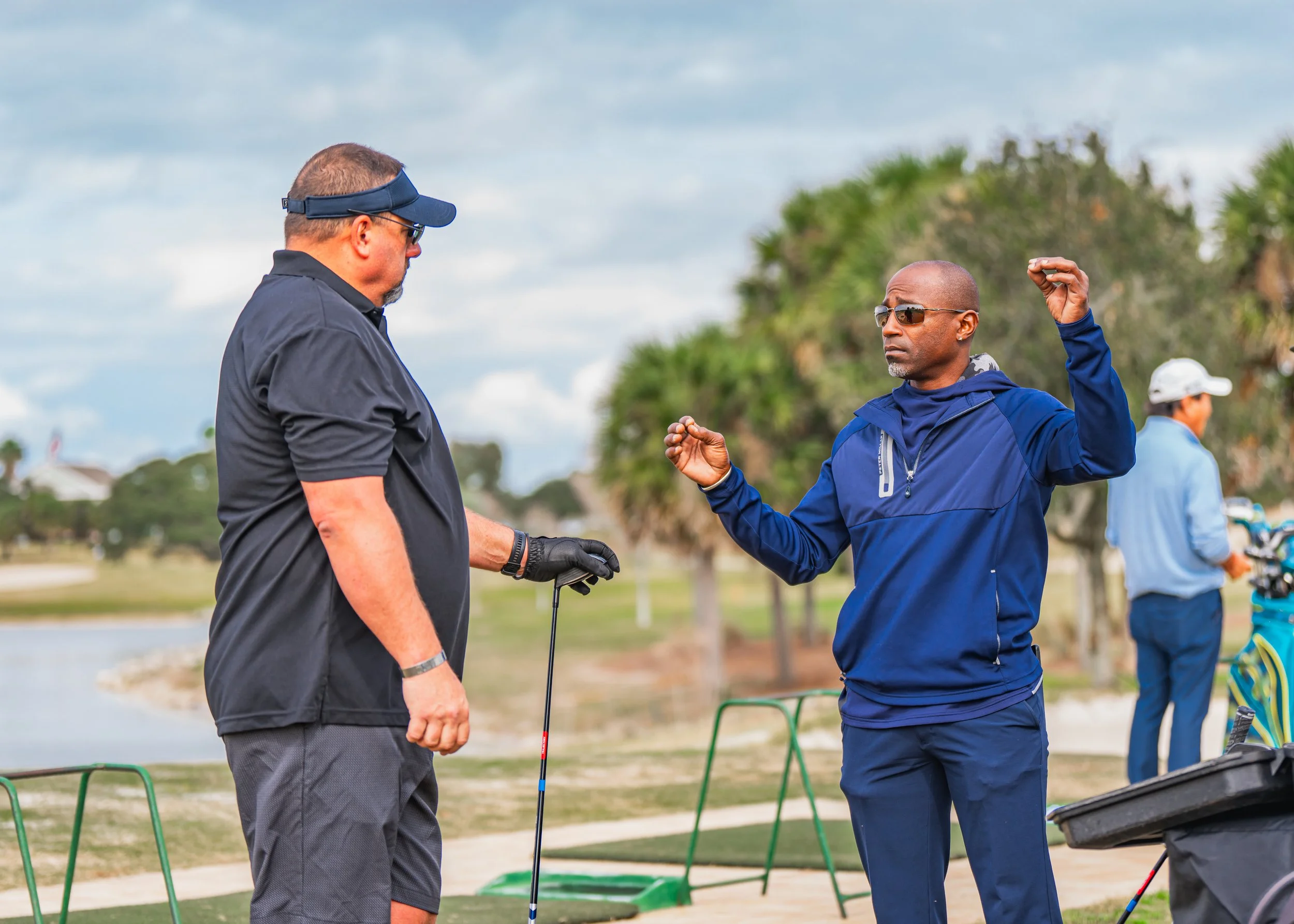Two men at a golf driving range, one is holding a golf club and wearing a black shirt, the other is in a blue jacket, sunglasses, and gesturing with his hands.