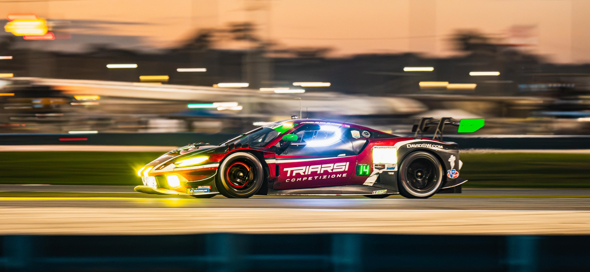Race car on track during sunset with motion blur, displaying branding and a bright front headlight.