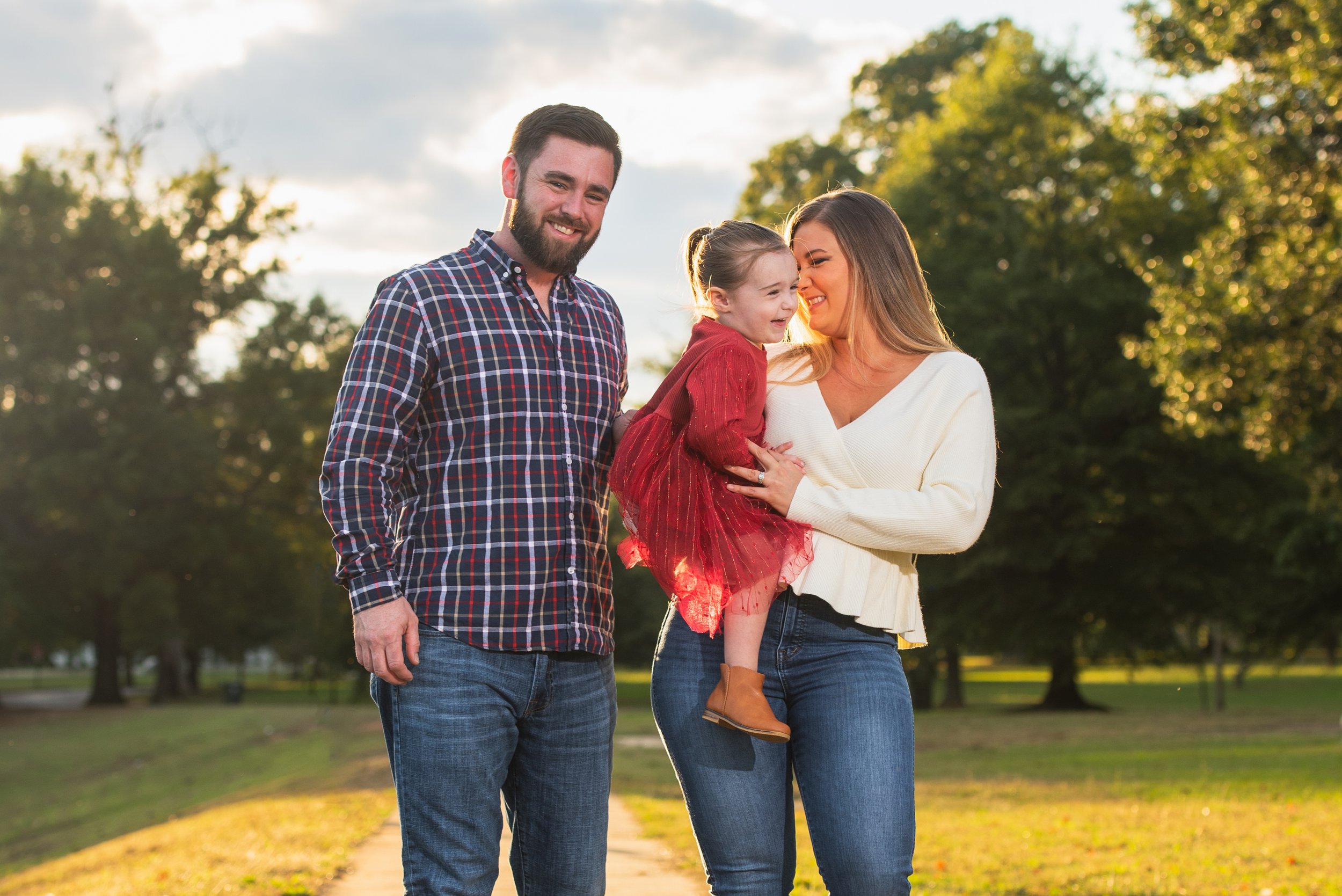 A family of three, a man, woman, and young girl, smiling and enjoying time outdoors in a park during the golden hour with trees and a pathway in the background.