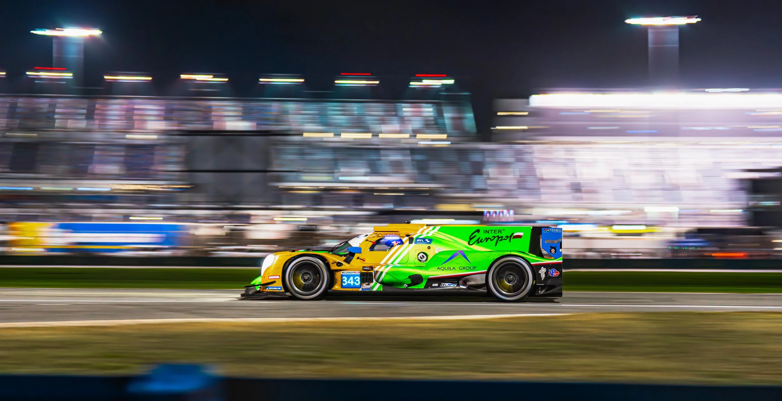 A brightly colored race car with the number 343 on its side speeding on a race track at night, with blurred background lights indicating high speed.