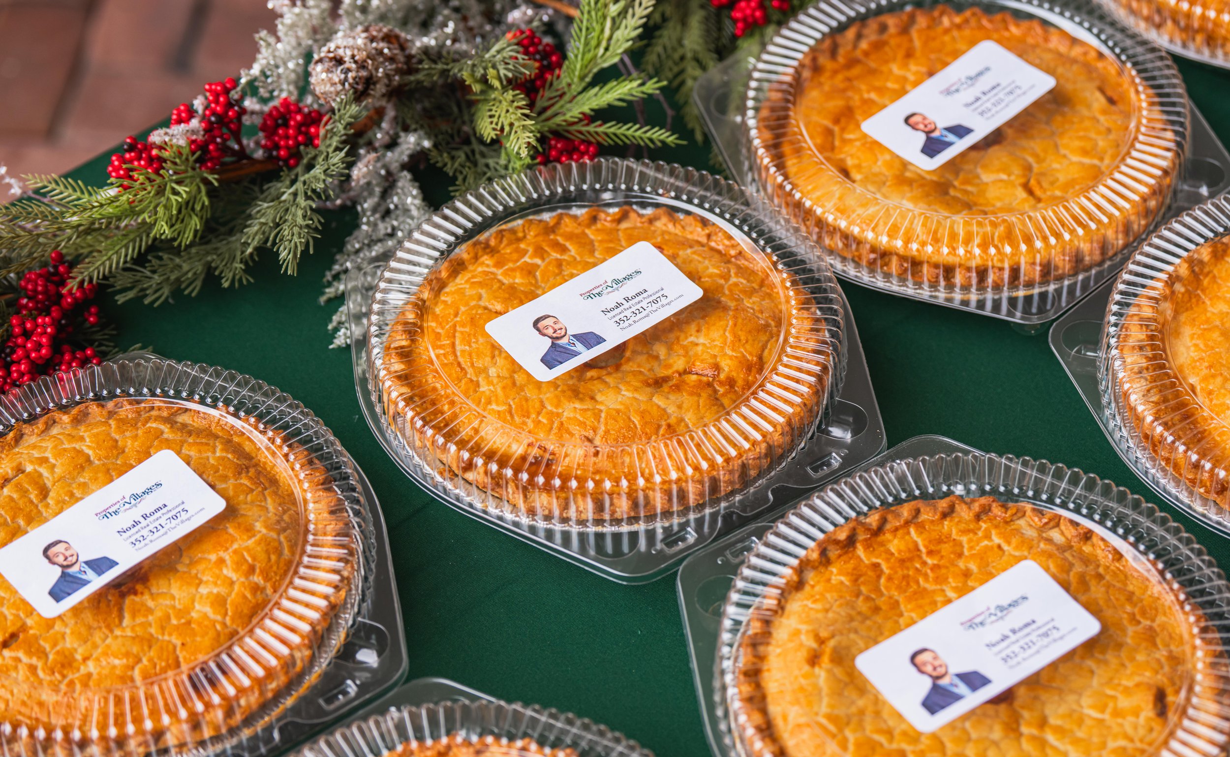 Multiple apple pies in clear plastic containers with labels, decorated with Christmas foliage and red berries on a green tablecloth.