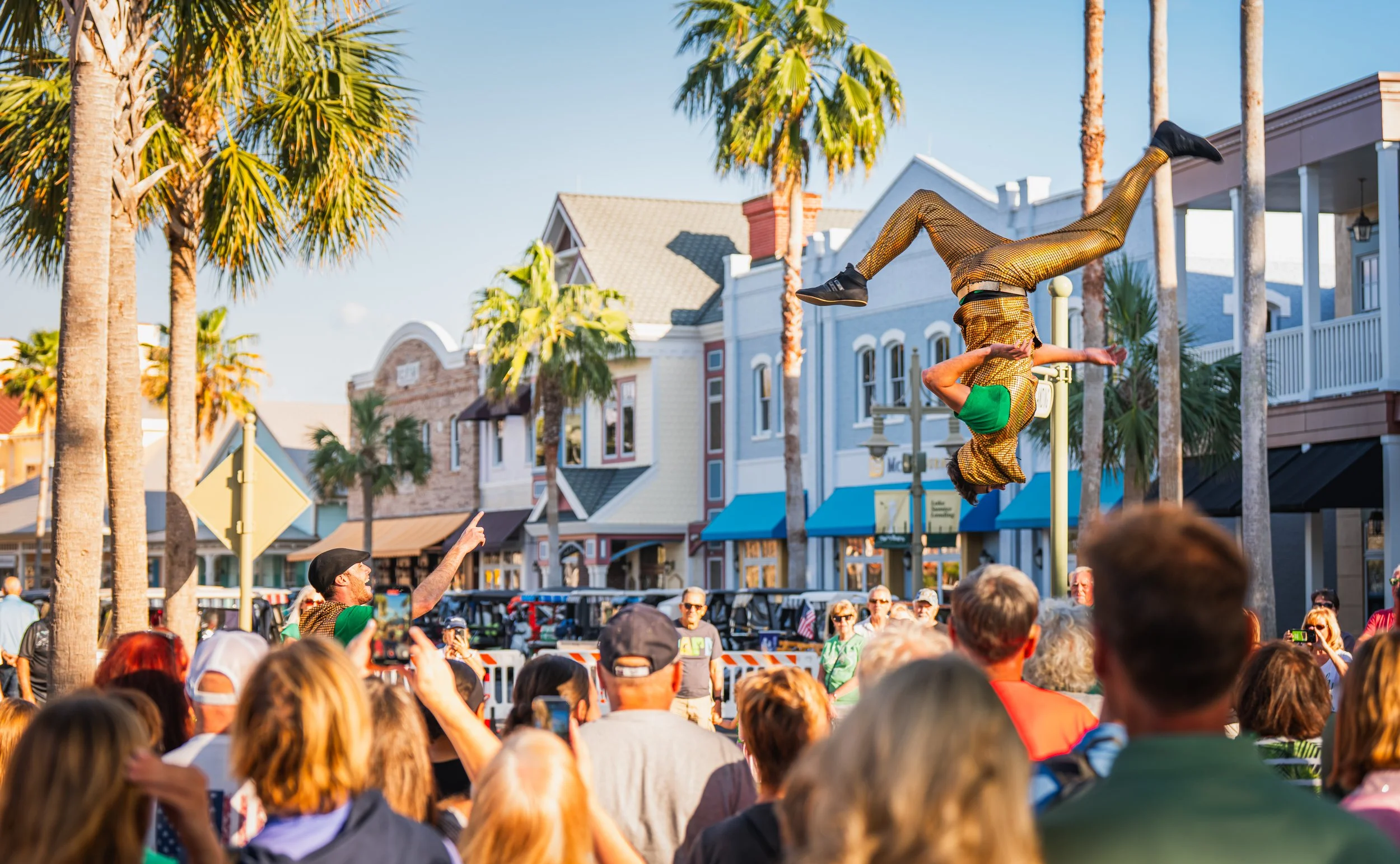 A street performer doing a backflip in front of a crowd on a sunny day with palm trees and colorful buildings.