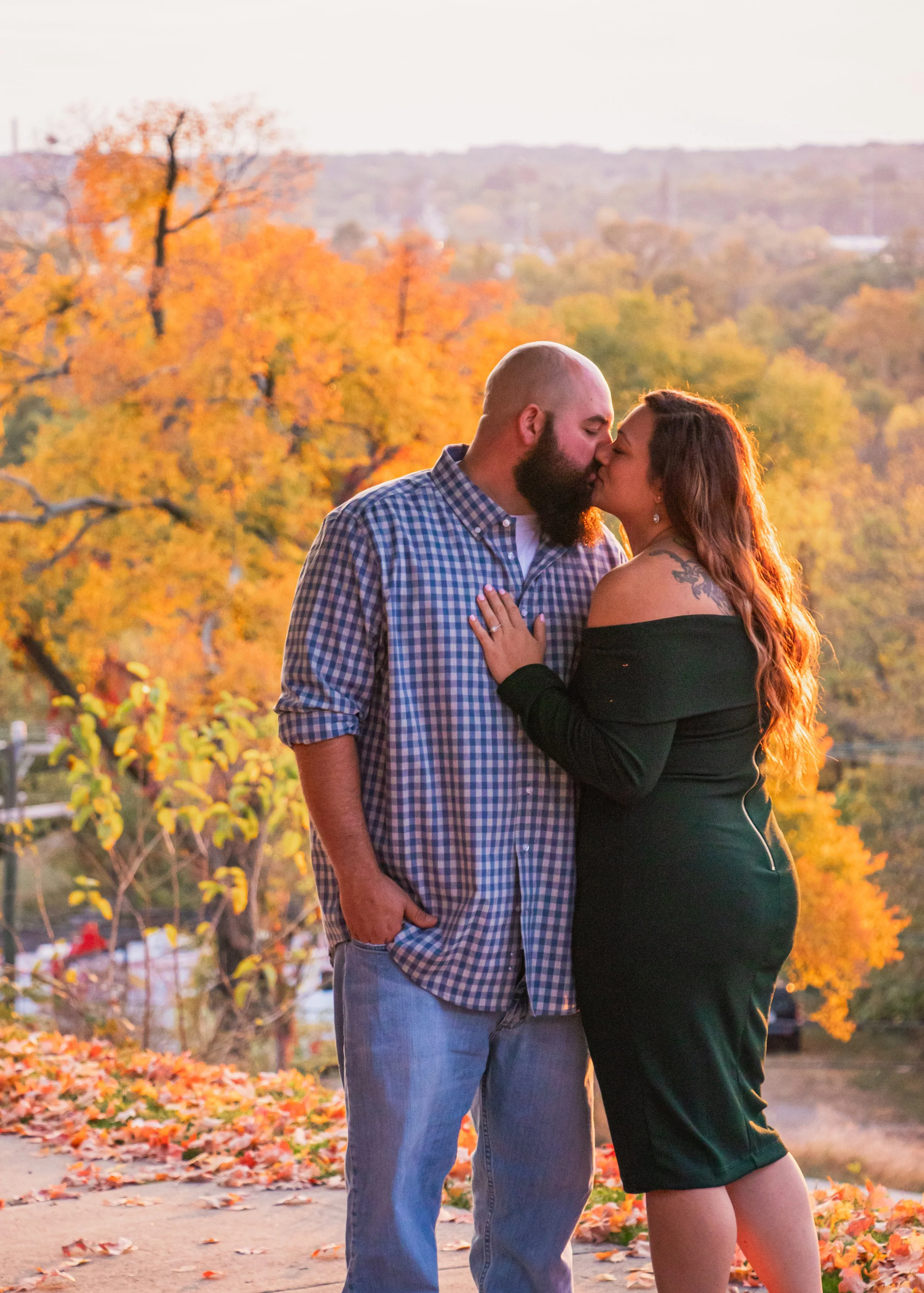 A couple sharing a kiss outdoors during fall, with colorful autumn trees and fallen leaves in the background.