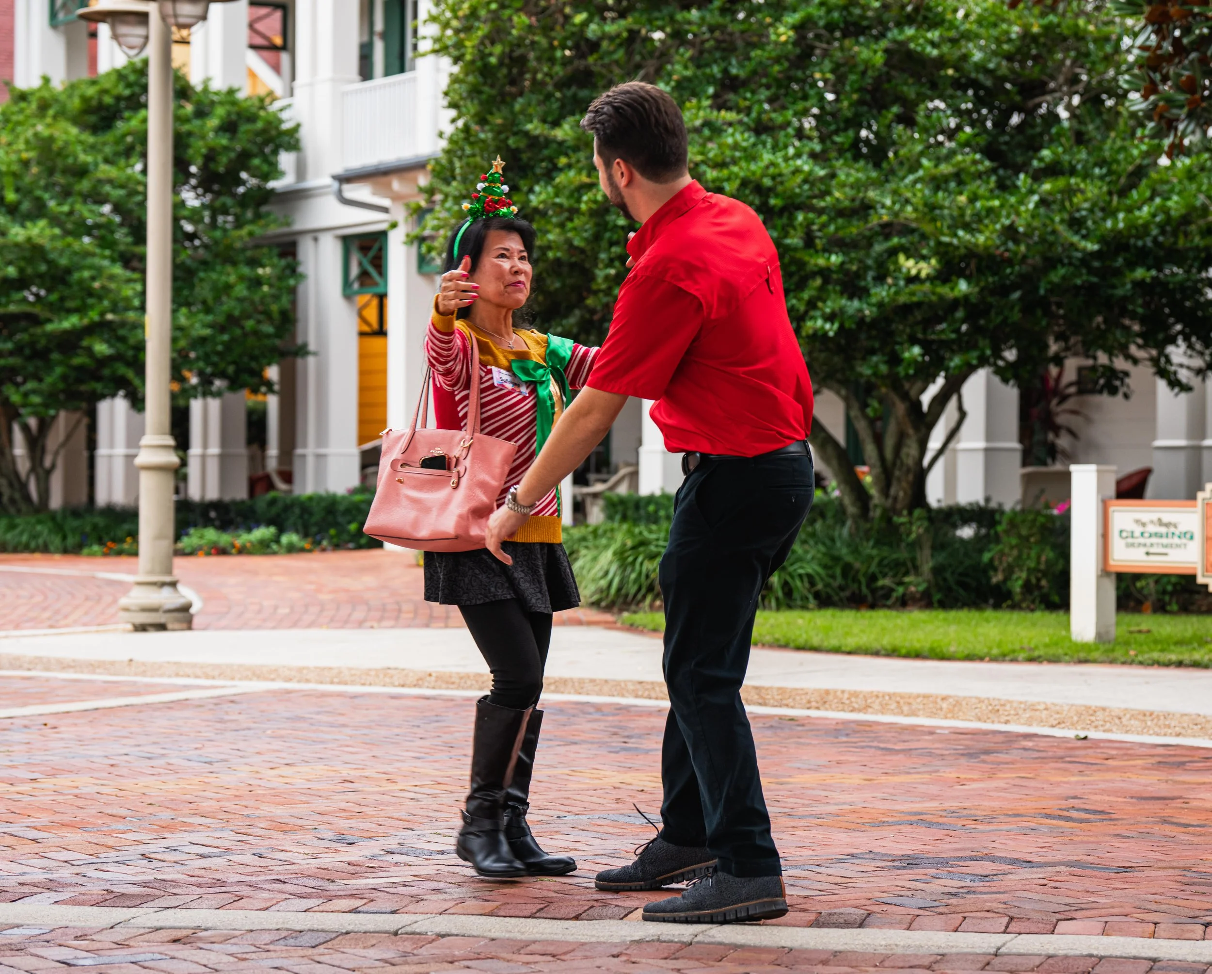 A woman wearing a Christmas tree headband, a red and white striped sweater, a green tie, and holding a pink purse, gestures to a man in a red shirt and black pants as they stand on a brick sidewalk outdoors in front of green trees and white buildings