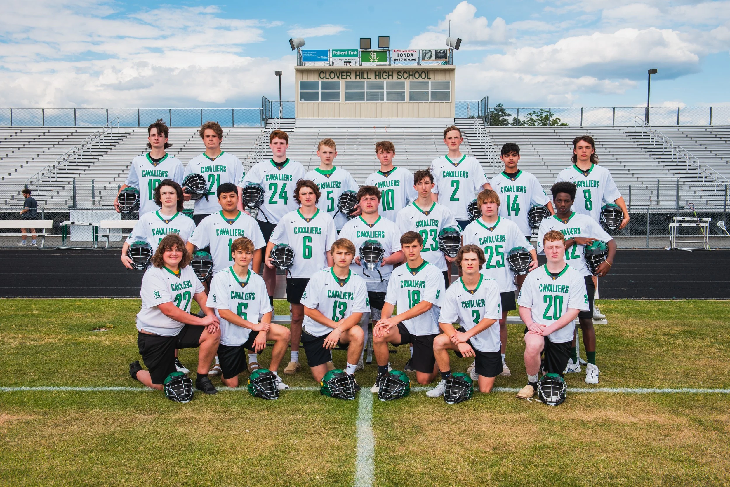 A high school football team poses for a team photo on a field with bleachers in the background. The team is wearing white jerseys with green lettering and black shorts, holding helmets, in front of a school stand that reads 'Clover Hill High School'.