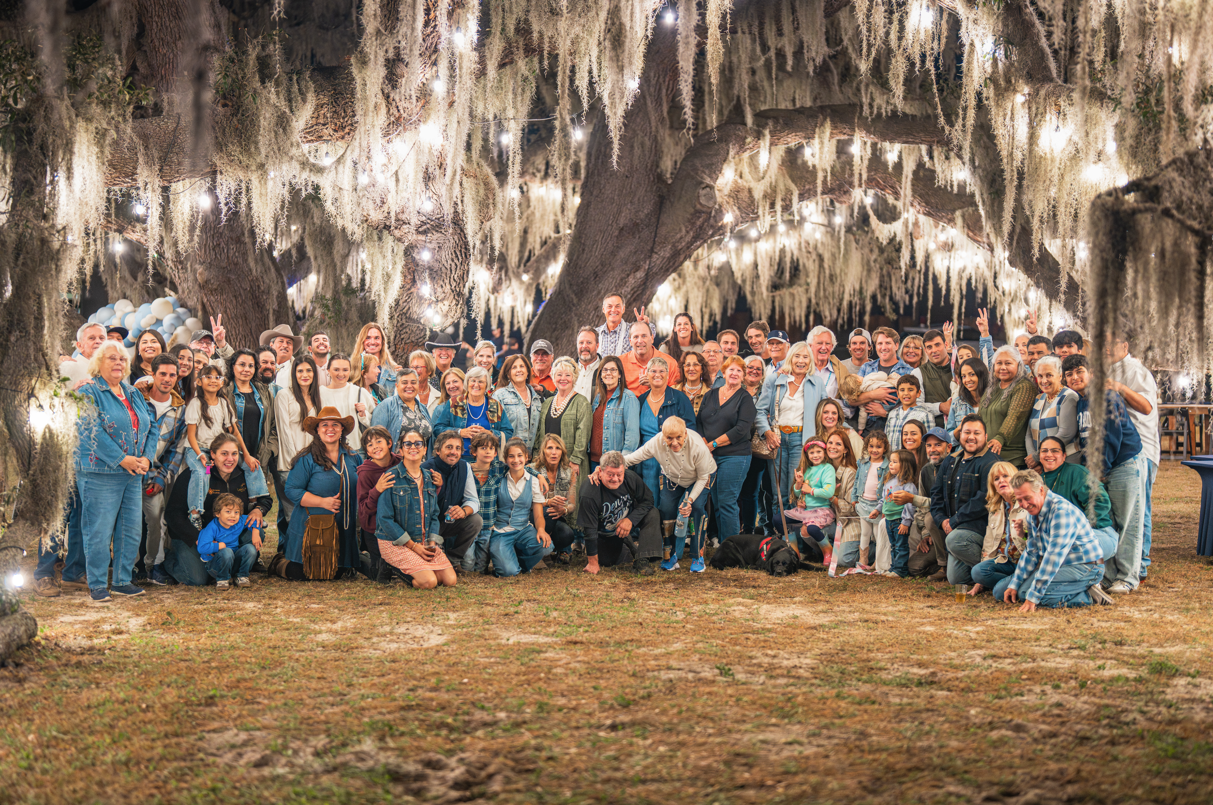 Large group of people, including children and adults, gathering outdoors at night under hanging moss and string lights, celebrating around a large tree with families and friends.