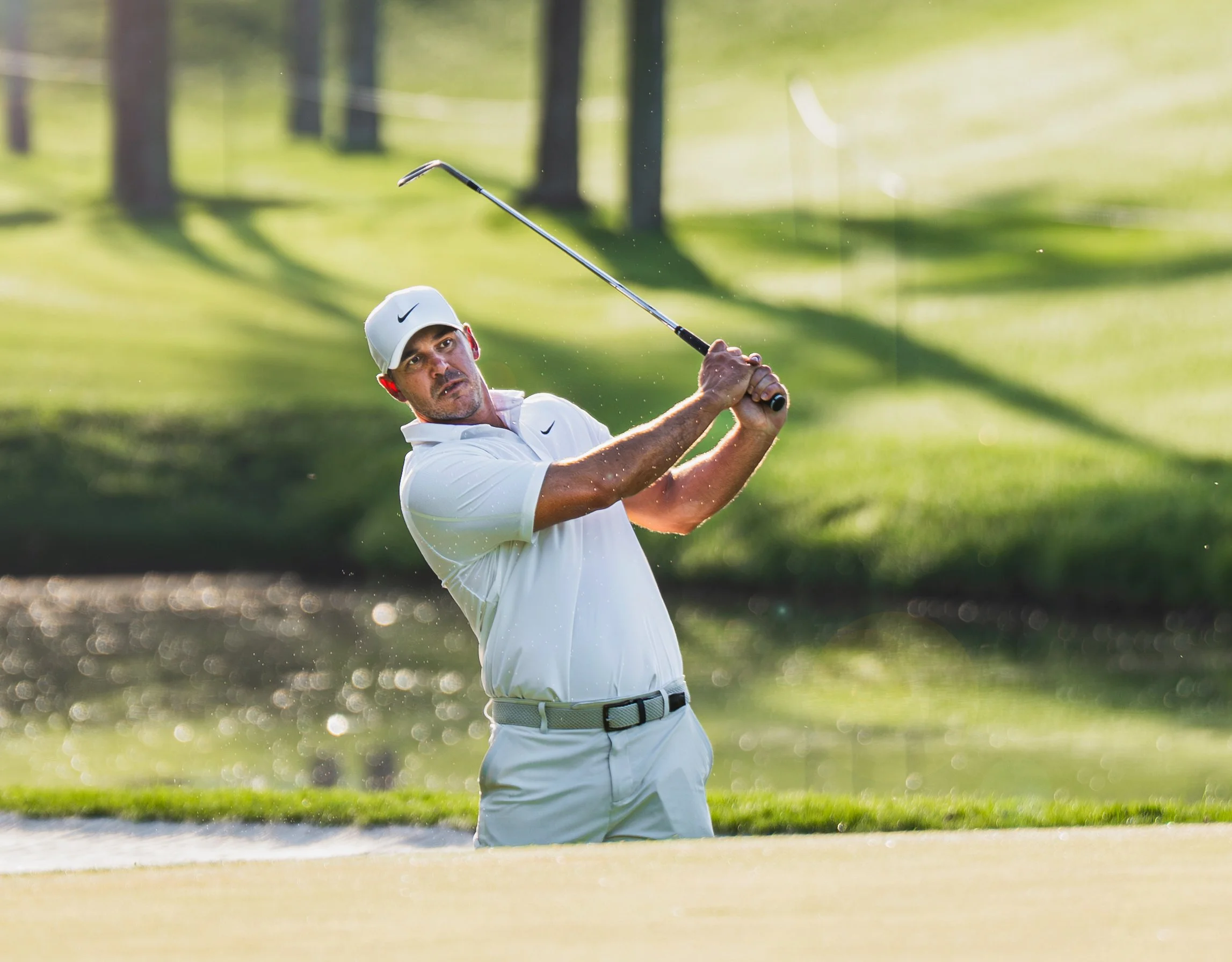 Brooks Koepka in a white shirt and cap swinging a golf club near a water hazard on a golf course.