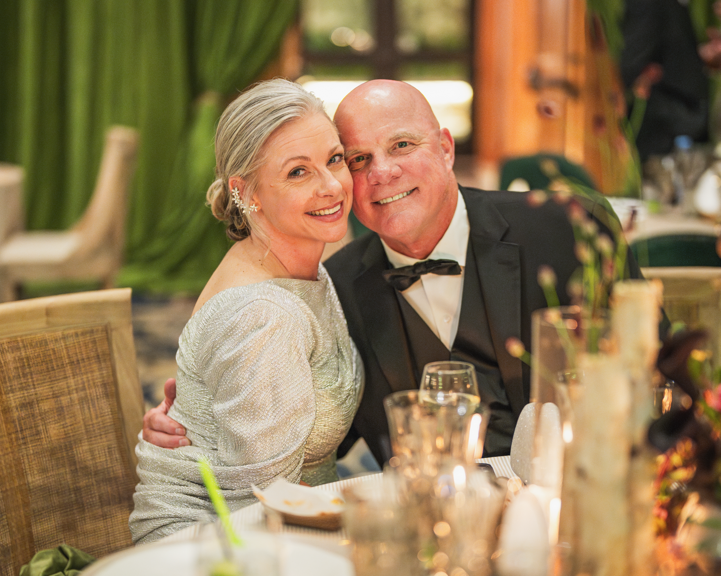 A smiling middle-aged couple dressed in formal attire, sitting at a decorated dinner table during a celebration or wedding reception.