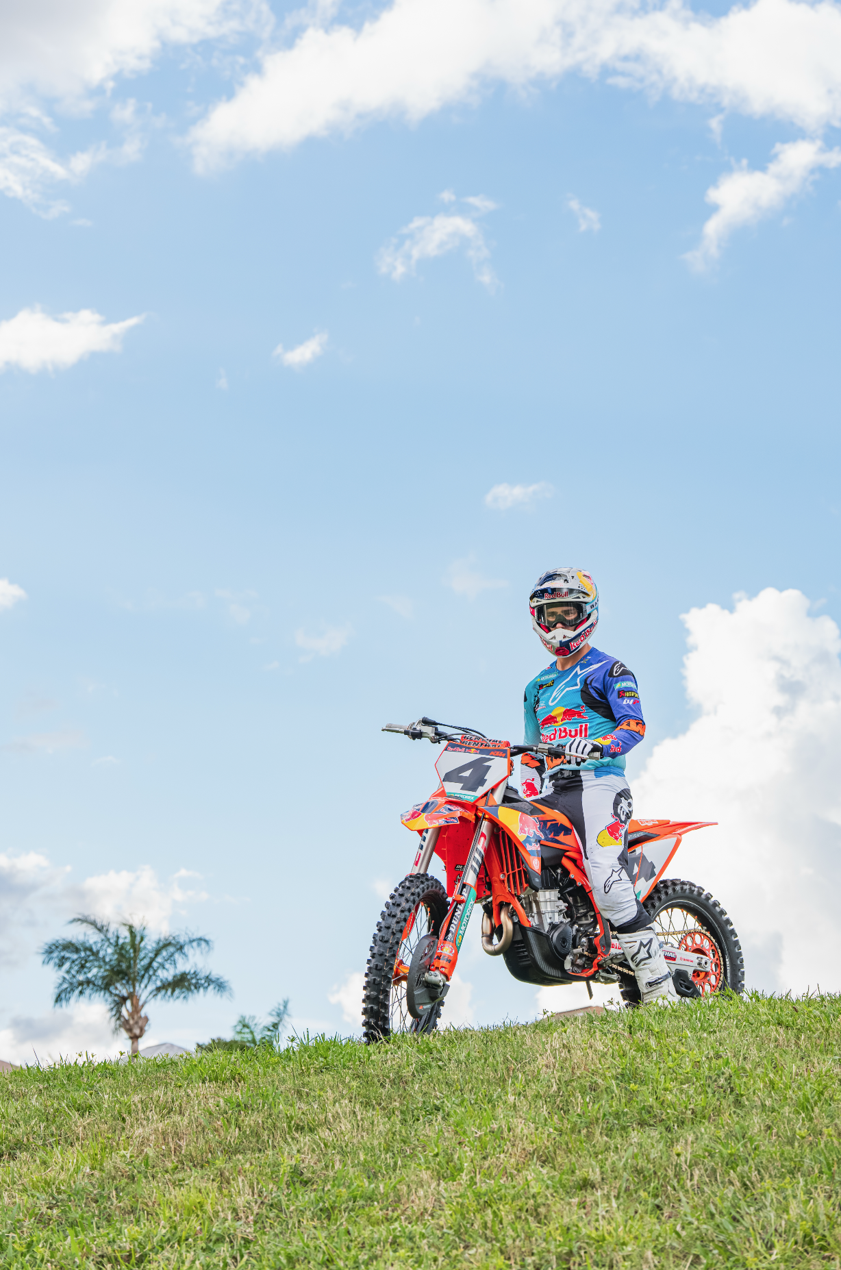 Motocross rider in blue and white gear sitting on orange dirt bike with number 4 on a grassy hill, under a blue sky with scattered clouds and a palm tree in the background.