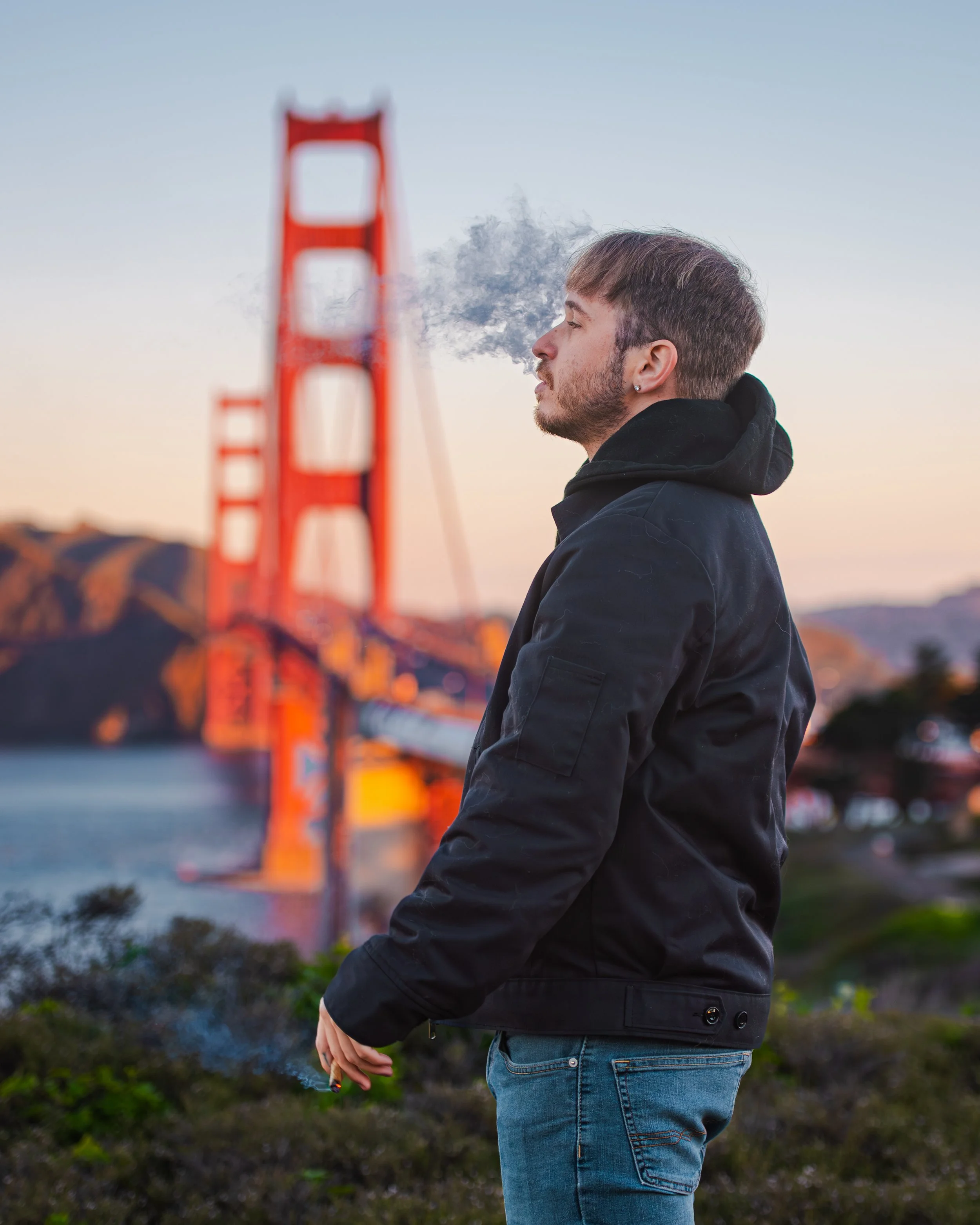 A young man in a black jacket and jeans smoking a cigarette with the Golden Gate Bridge in San Francisco in the background during sunset.