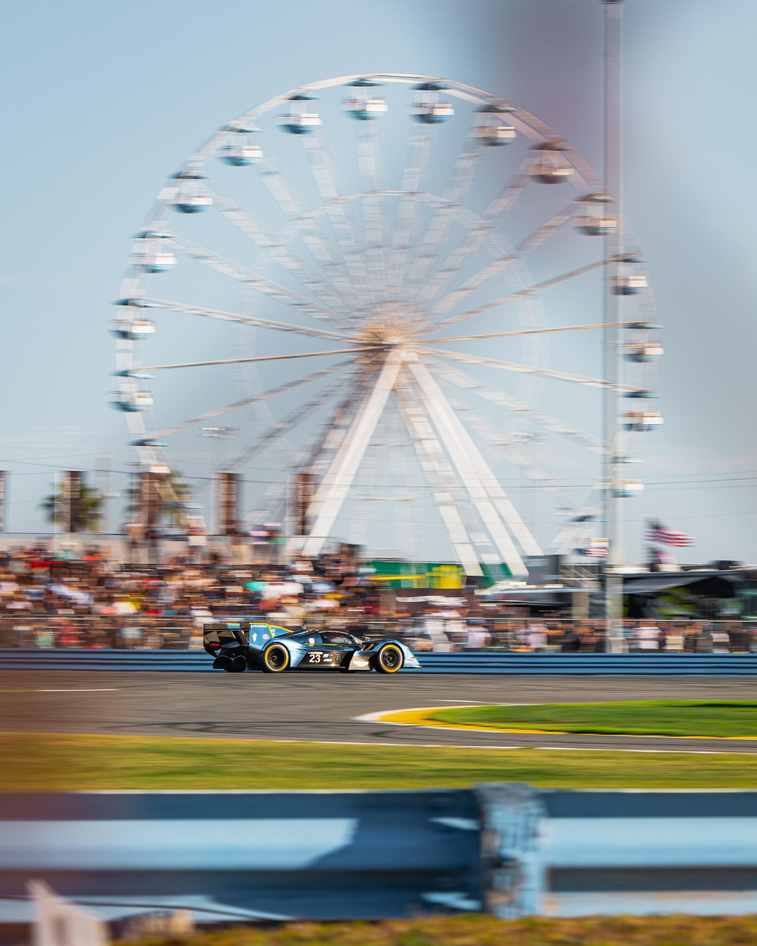A race car driving on a racetrack with a large Ferris wheel in the background and a crowd of spectators watching.