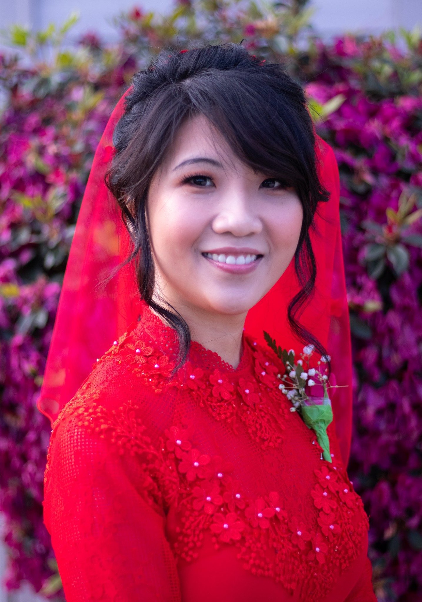 A smiling bride dressed in a red traditional dress with intricate embroidery and a floral corsage, standing outdoors in front of vibrant pink and purple flowers.