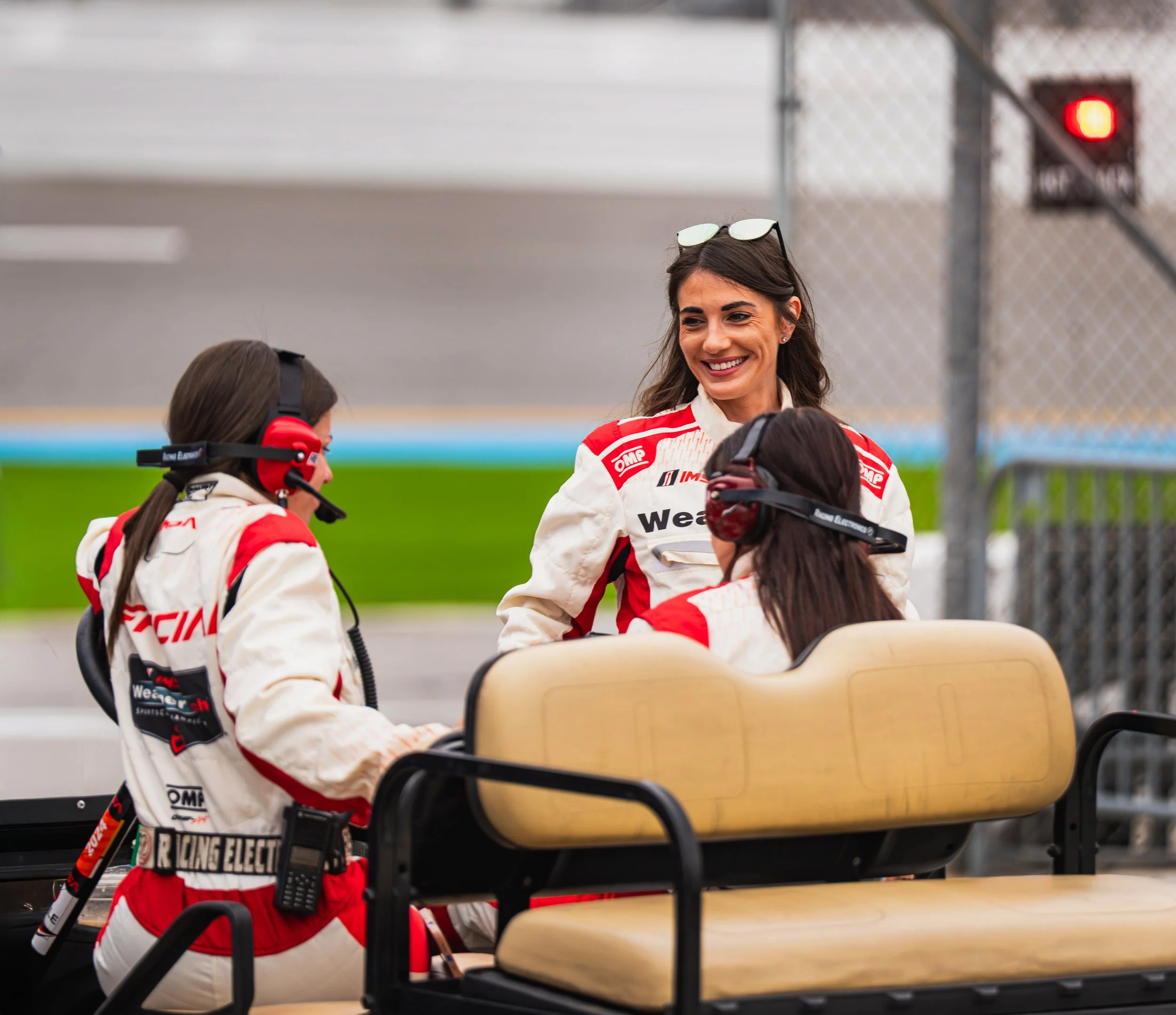 Women in racing suits smiling and talking near a medical golf cart at a racetrack.