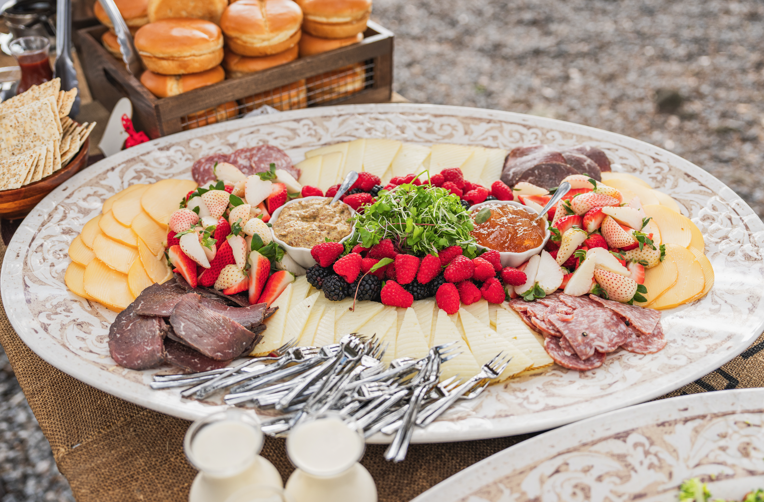 Cheese platter with sliced cheeses, fresh strawberries, raspberries, blackberries, and microgreens, accompanied by jams and meats, on an ornate serving tray at a buffet table.