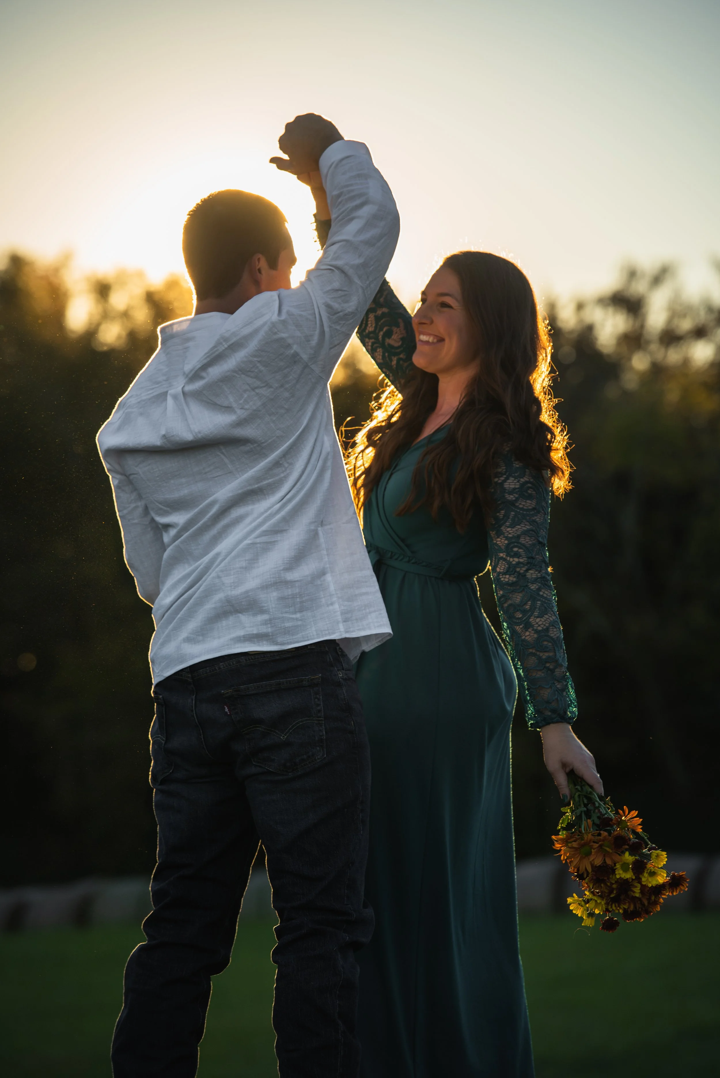 A couple sharing a tender moment outdoors during sunset, with the woman holding a bouquet of flowers and smiling at the man who is raising his hand towards her.