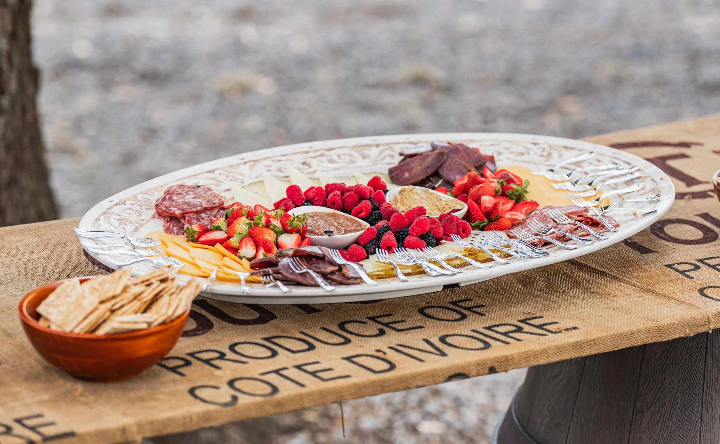 Charcuterie board with assorted cheeses, strawberries, raspberries, blackberries, sliced meats, and a dipping sauce, alongside a bowl of crackers on a rustic table.