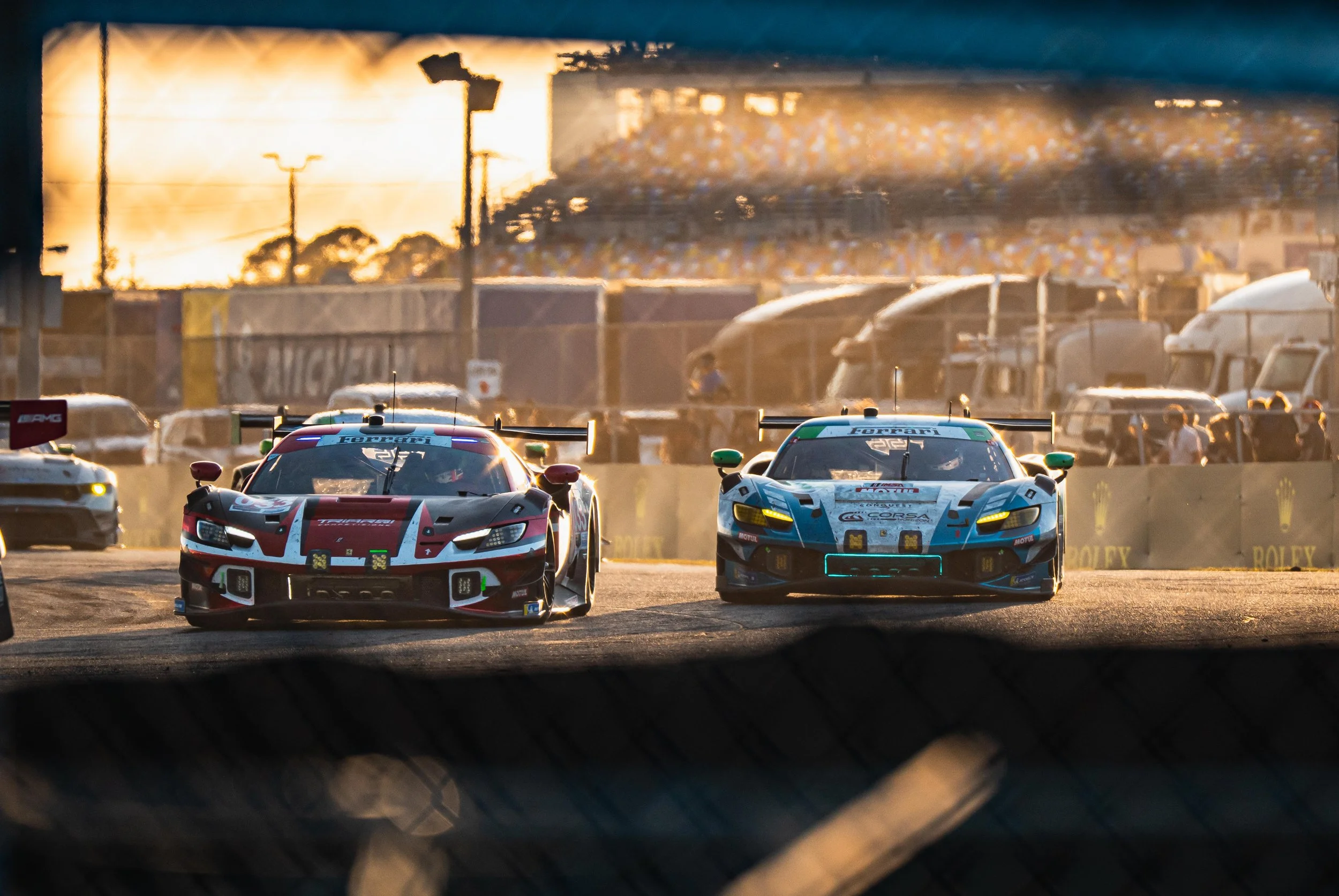 Two race cars on a race track during sunset, with a background of trucks, tents, and spectators.