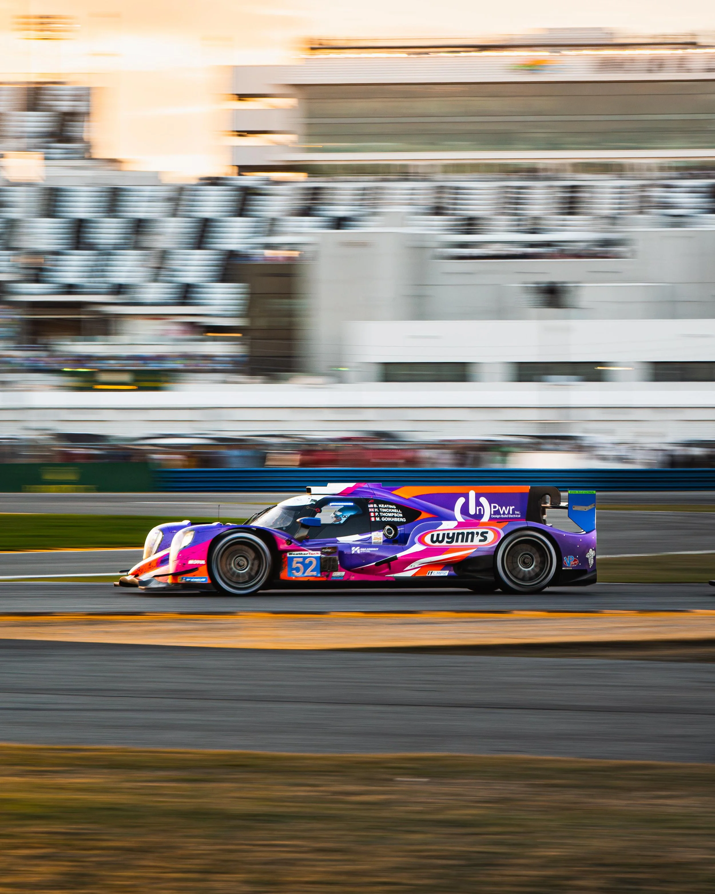A race car with purple, pink, and orange colors speeds along a track, with blurred background of grandstands and buildings.