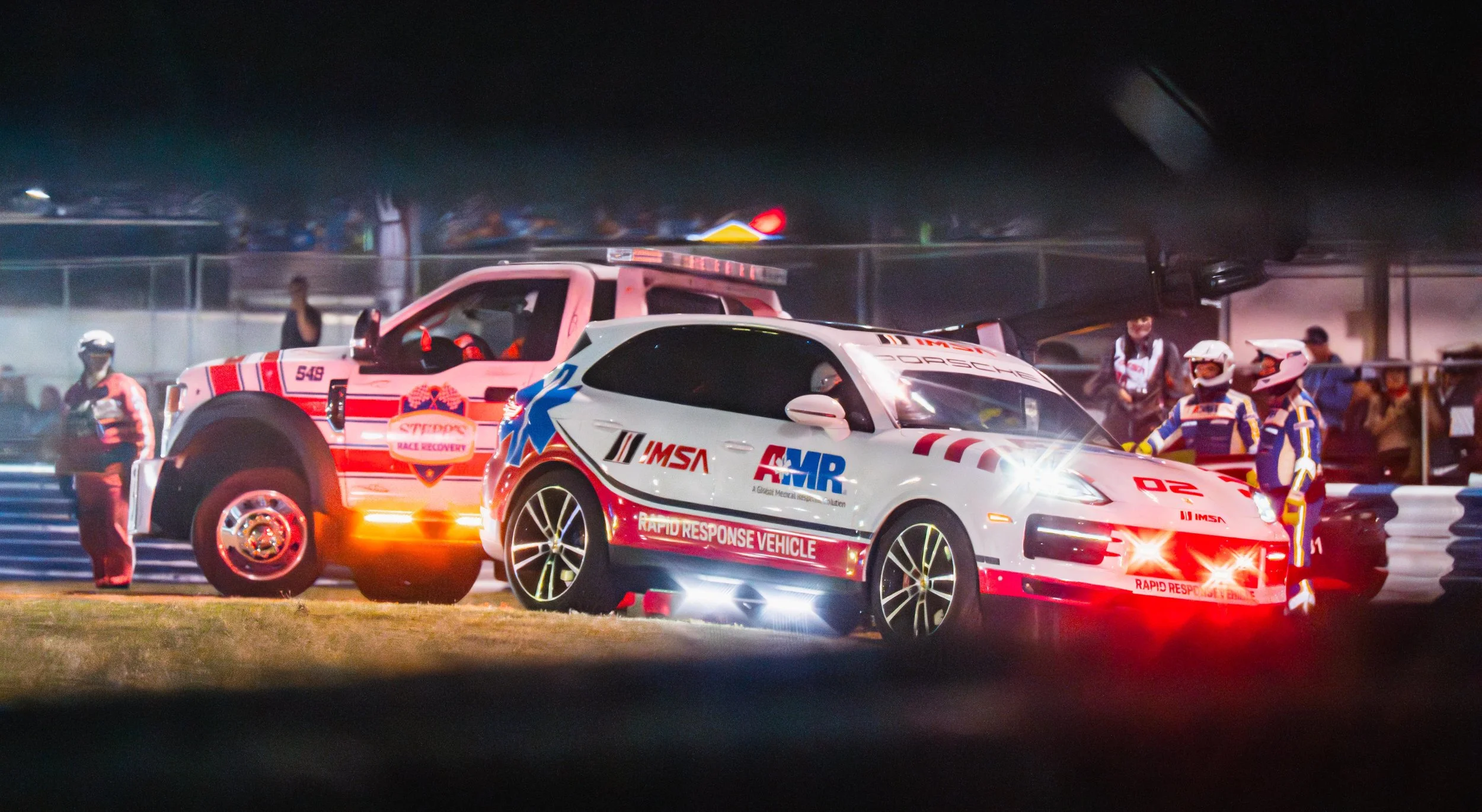 Race car and emergency response vehicle on a race track at night with crew members and race officials nearby.