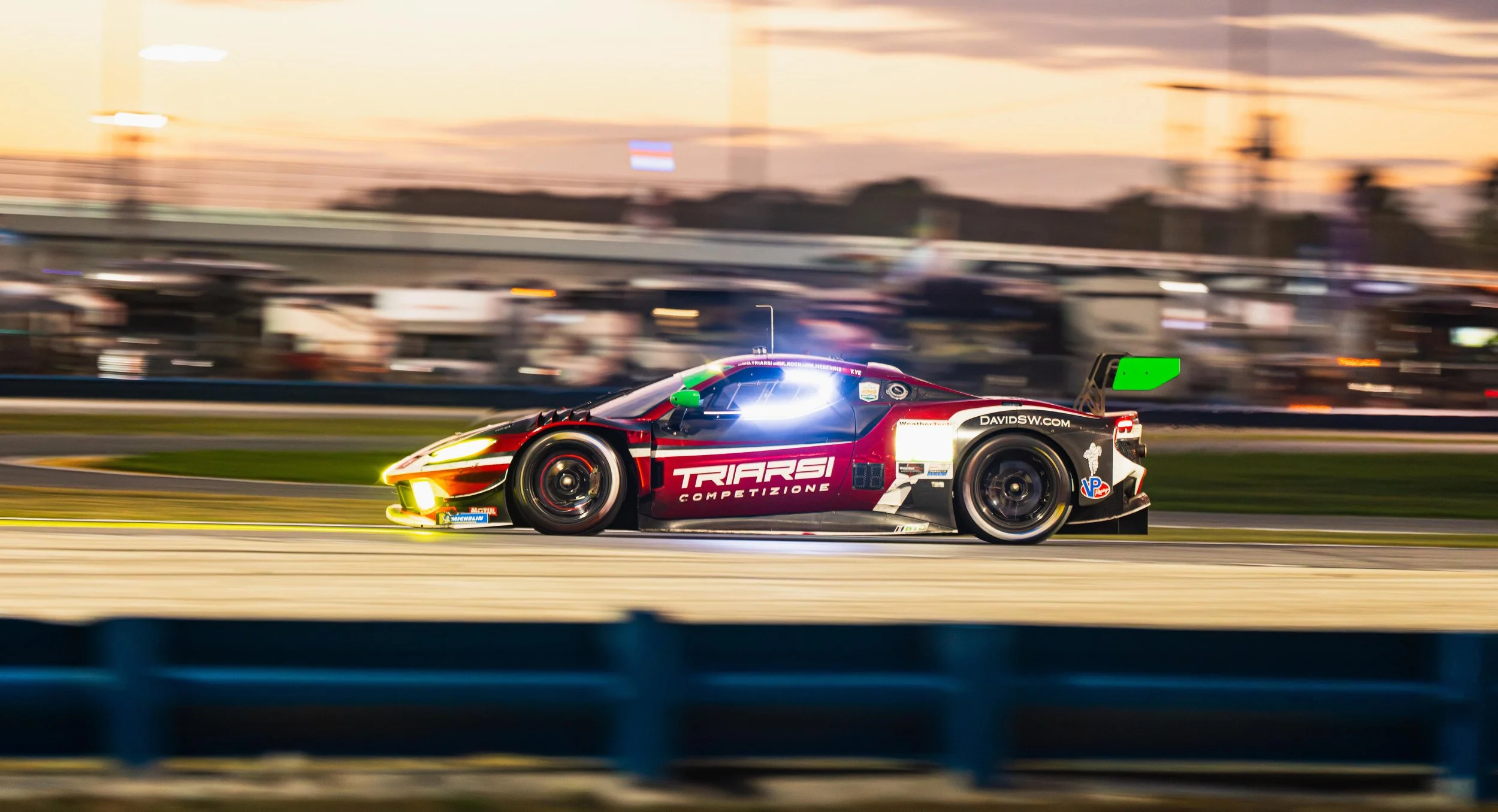 Race car speeding on a racetrack at sunset, branded with 'TRIARSI COMPETIZIONE' and various sponsor logos.