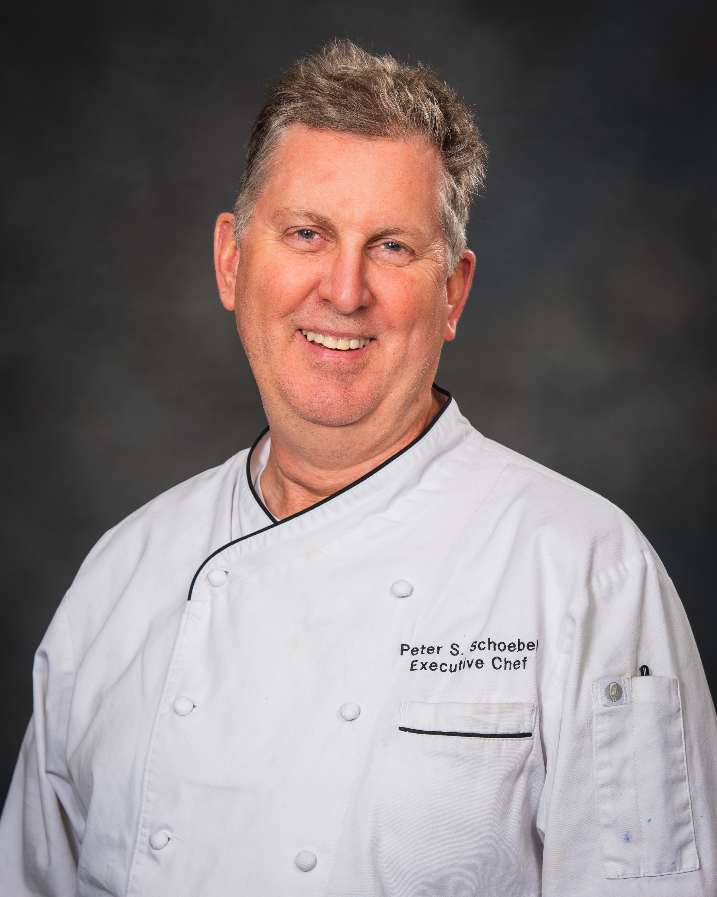 Portrait photography, Central Florida. Corporate headshots. A smiling man in a white chef's coat with embroidered text: Peter S. Schoebel, Executive Chef, against a dark background.