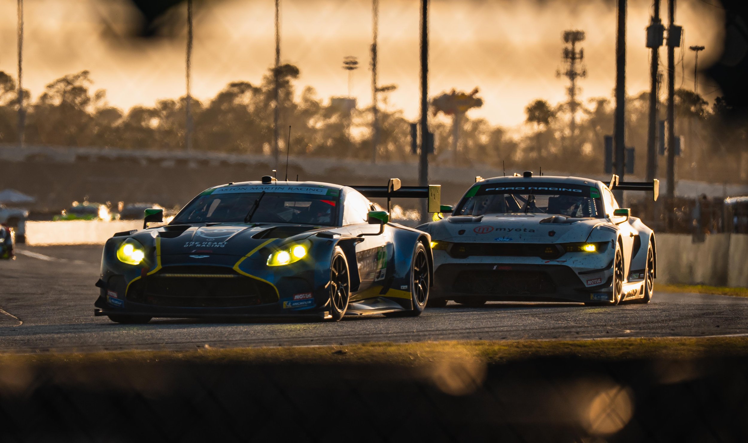 Two race cars on a track, chasing each other during sunset with trees and power lines in the background.