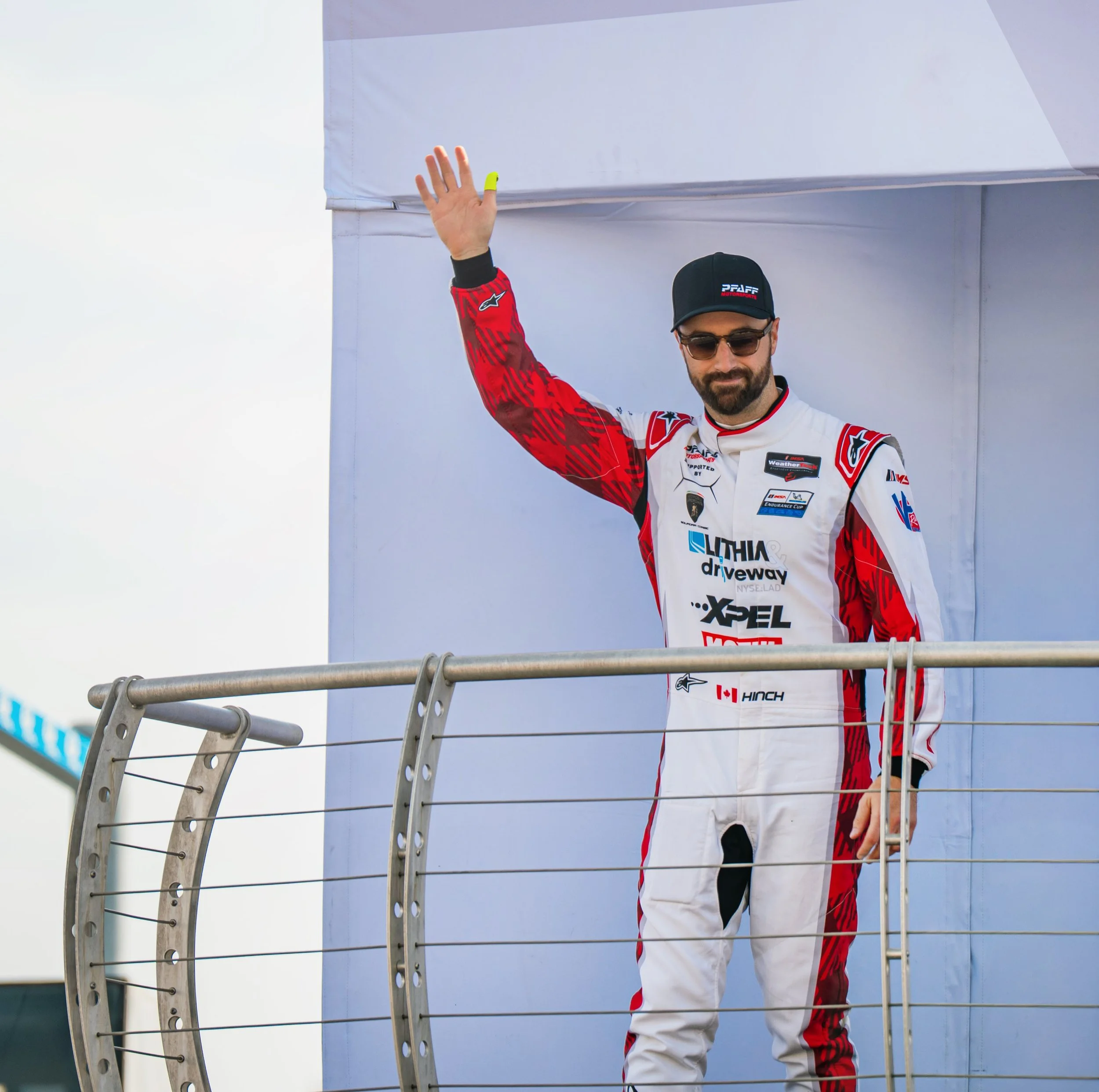 A race car driver in a white and red racing suit waving from the podium, wearing sunglasses and a black cap, with a railing in front and a large white backdrop behind him.