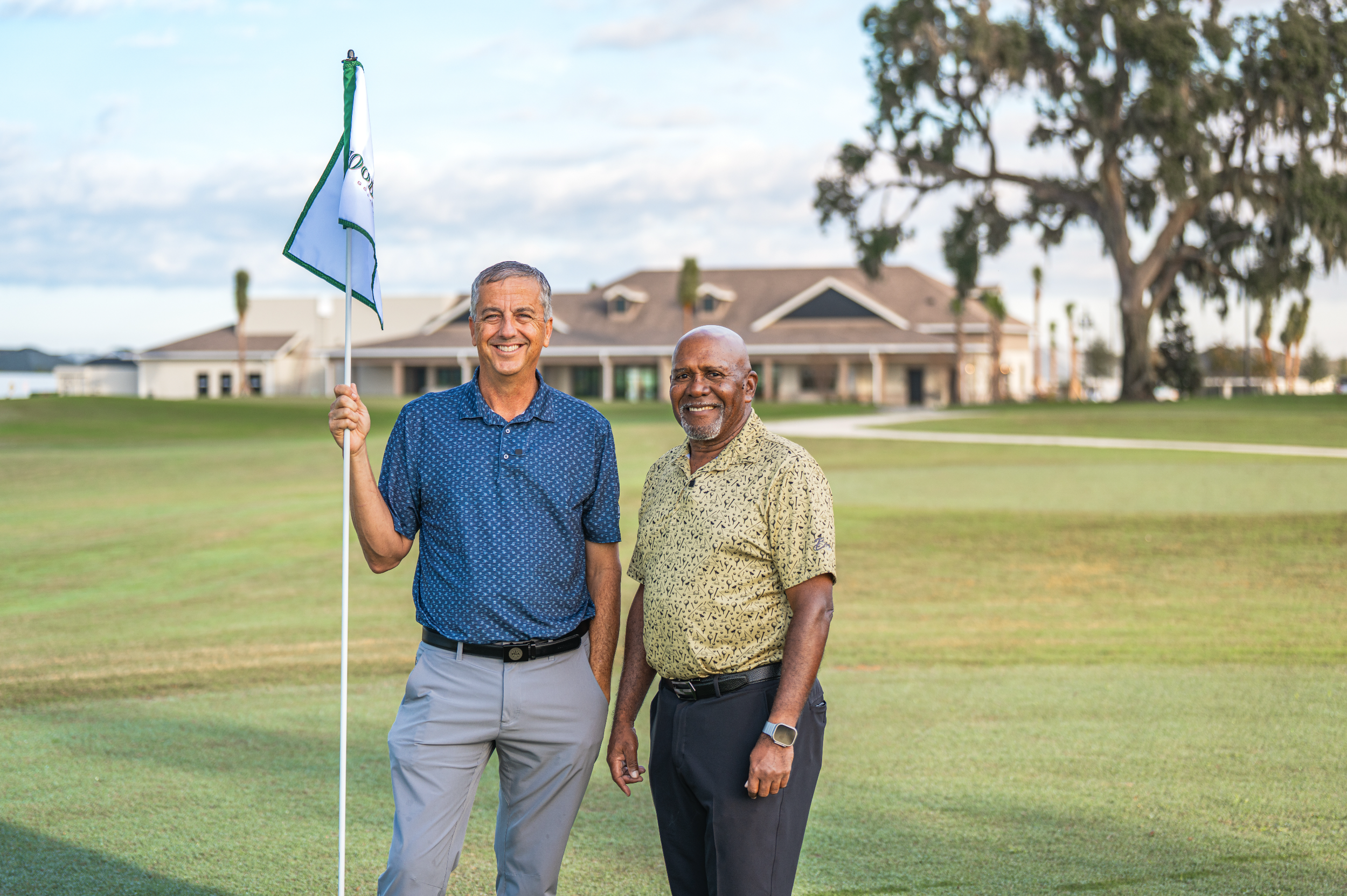 Two men standing on a golf course with a clubhouse in the background. One man is holding a golf flag and both are smiling. The scene is outdoors during daytime.