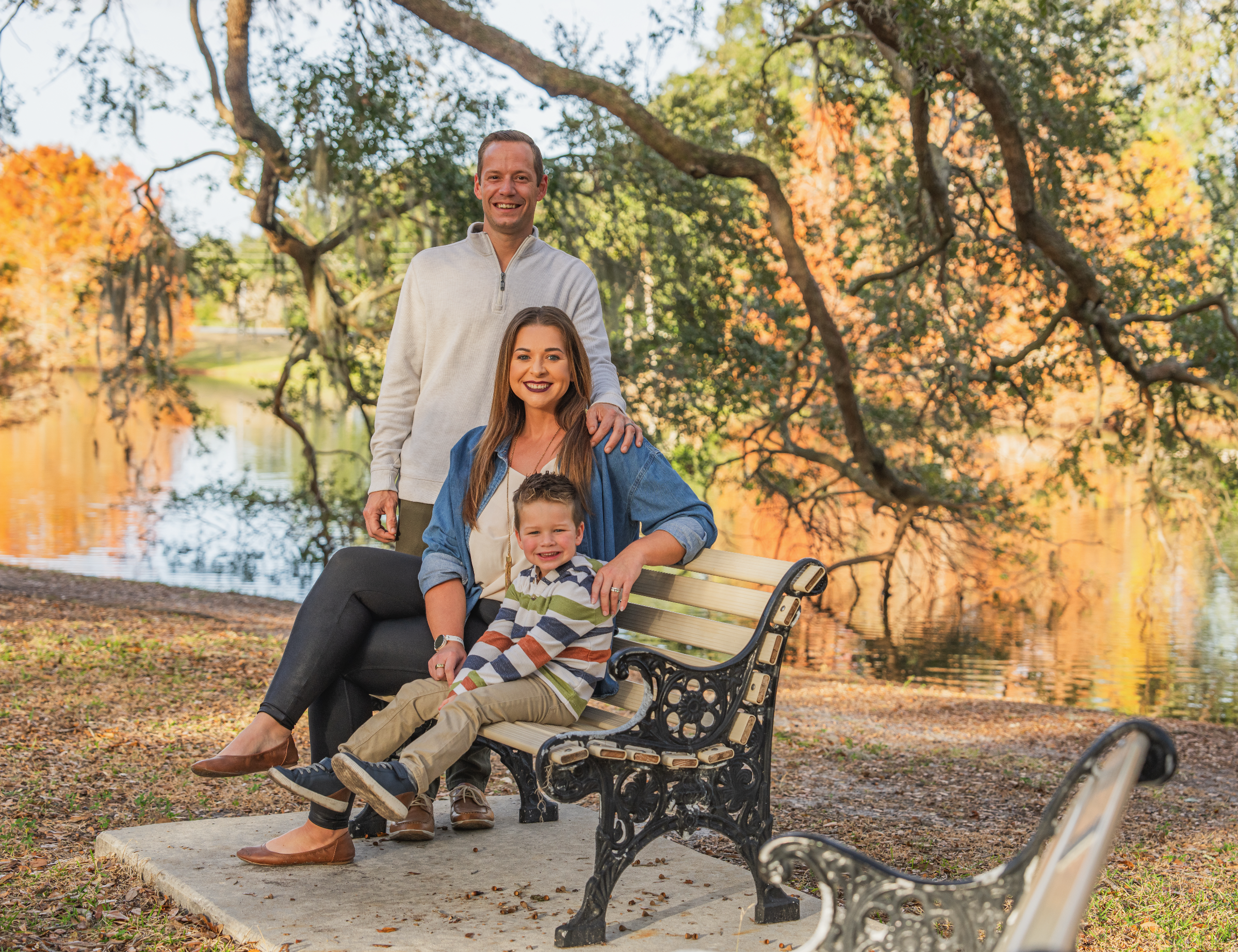 A happy family of three in autumn attire outdoors by a lake, with colorful fall trees in the background. The woman is sitting on a park bench, the young boy is sitting on her lap, and the man stands beside them with a hand on her shoulder.