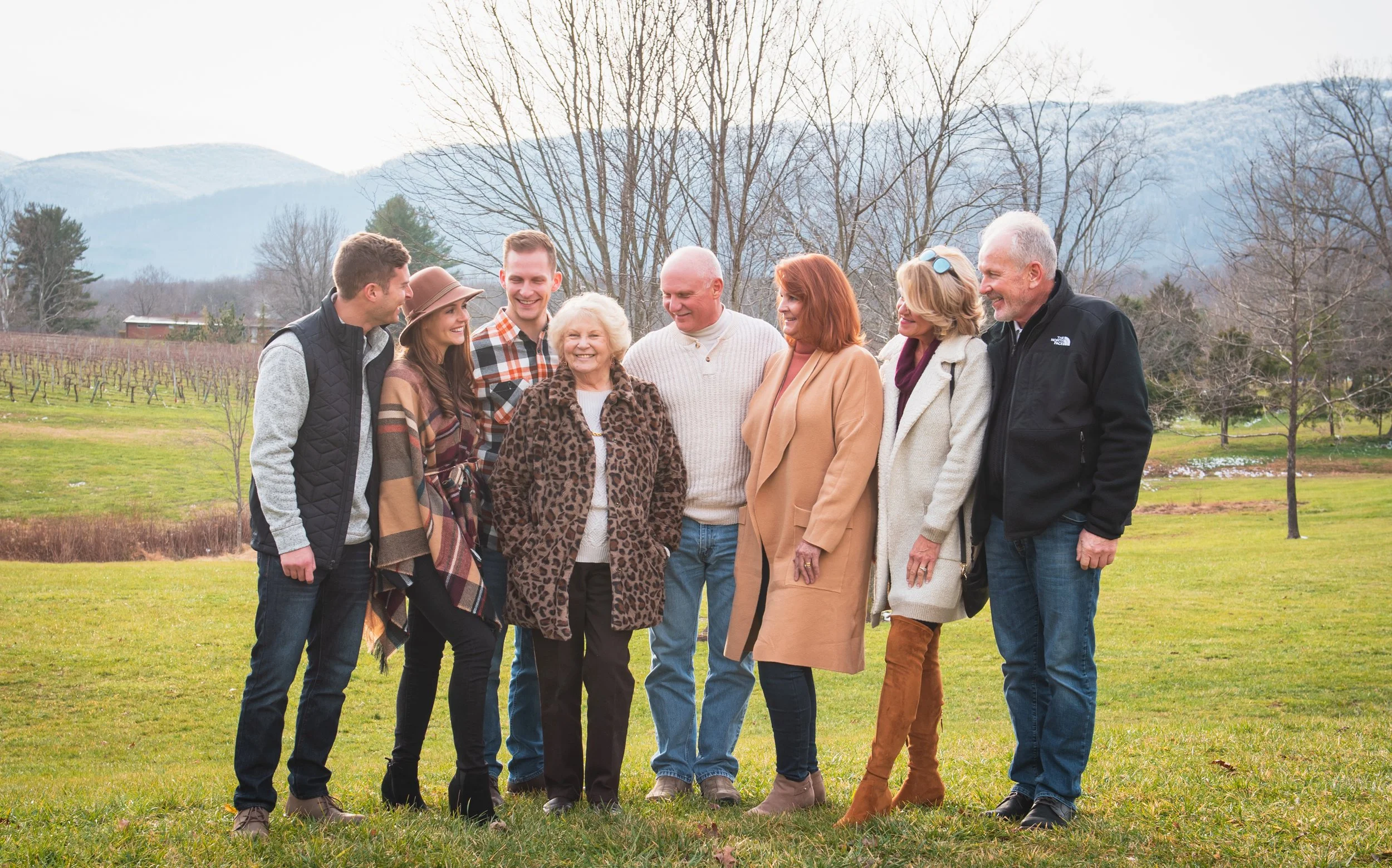 A family of nine people standing outdoors on a grassy field, smiling and engaging in conversation, with mountains and leafless trees in the background during an overcast daytime.