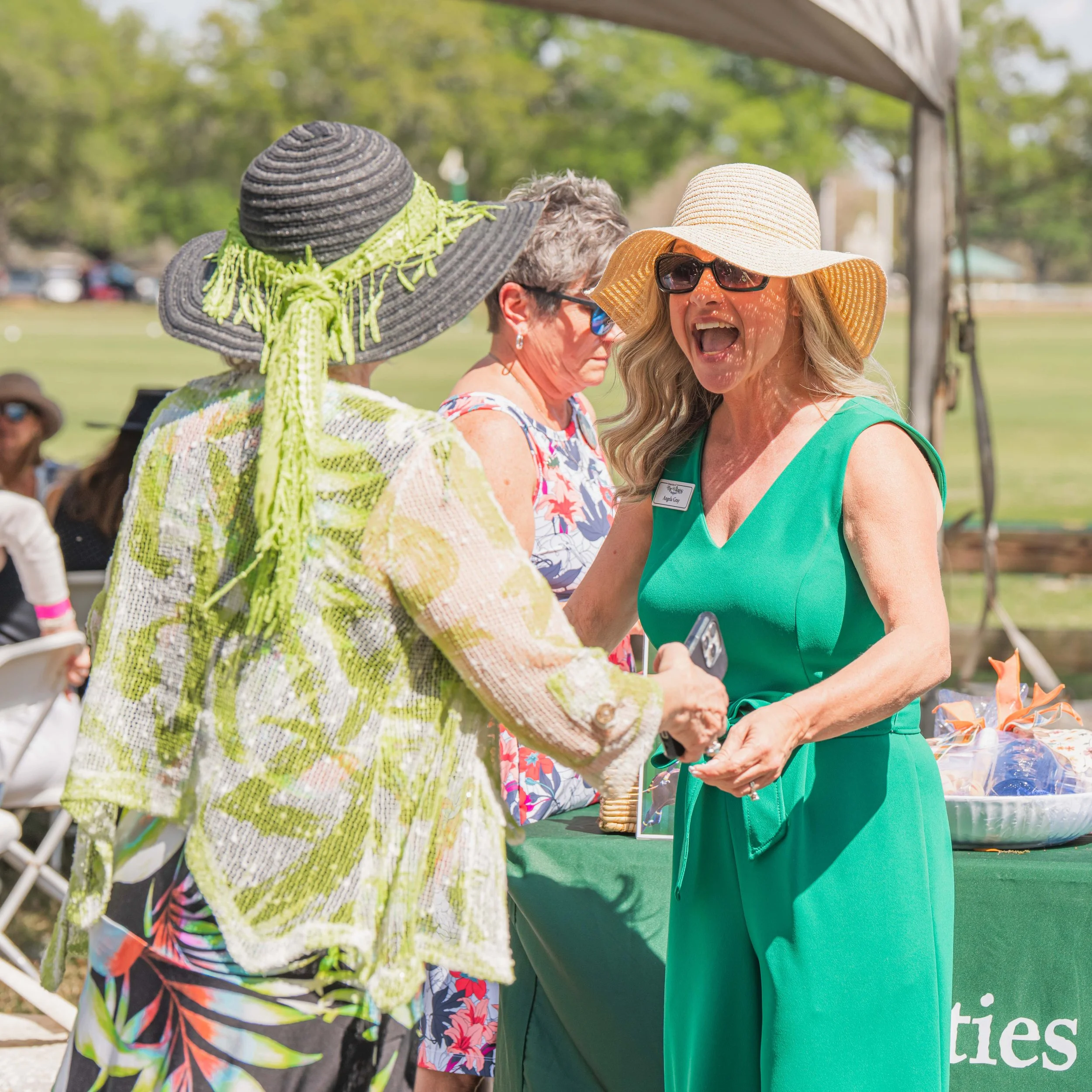 Women conversing outdoors at a sunny event, one wearing a large hat with a bow, sunglasses, and a green dress, others in colorful attire, under a canopy with trees in the background.