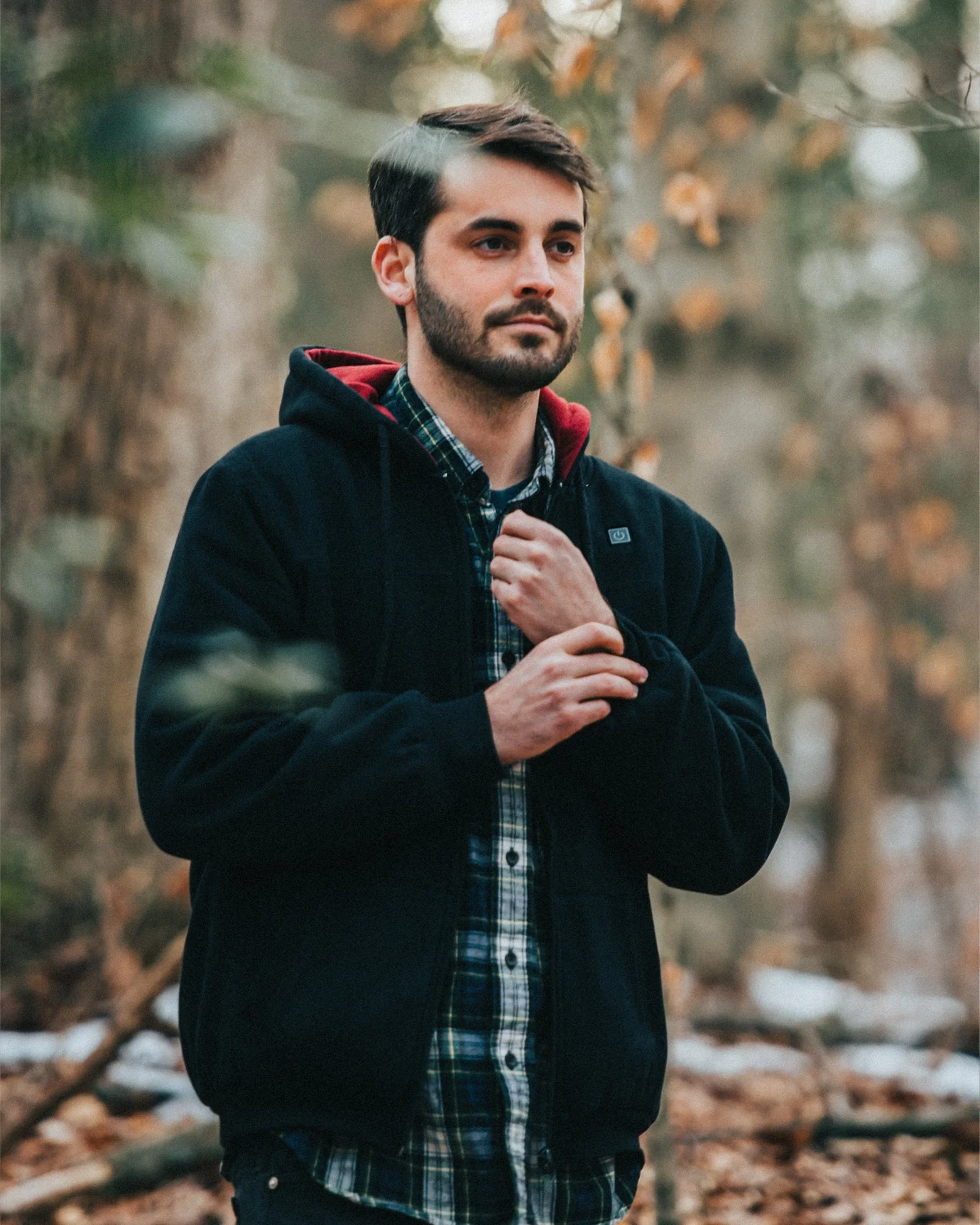 A man with dark hair and a beard standing outdoors in a forest, wearing a black jacket with a hood and a plaid shirt, looking contemplative.